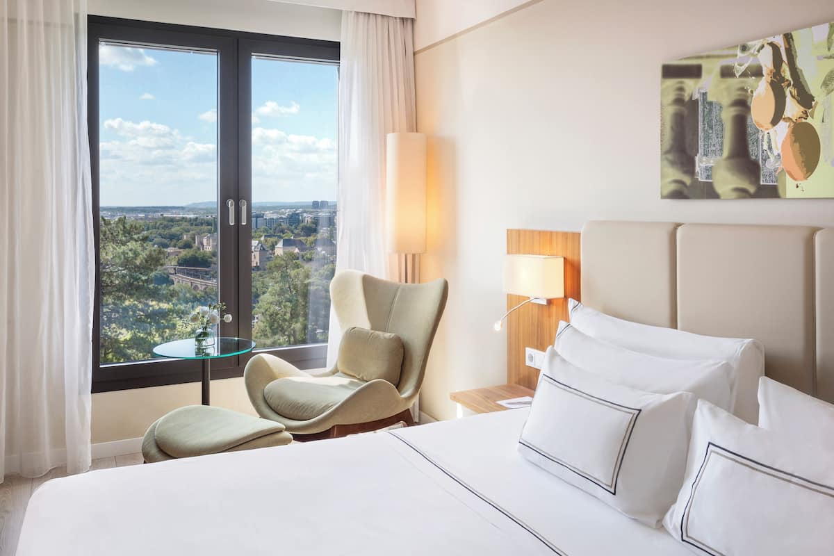Habitación de hotel con una cama grande, una silla, una mesita, una lámpara y vista a la ciudad desde la ventana.