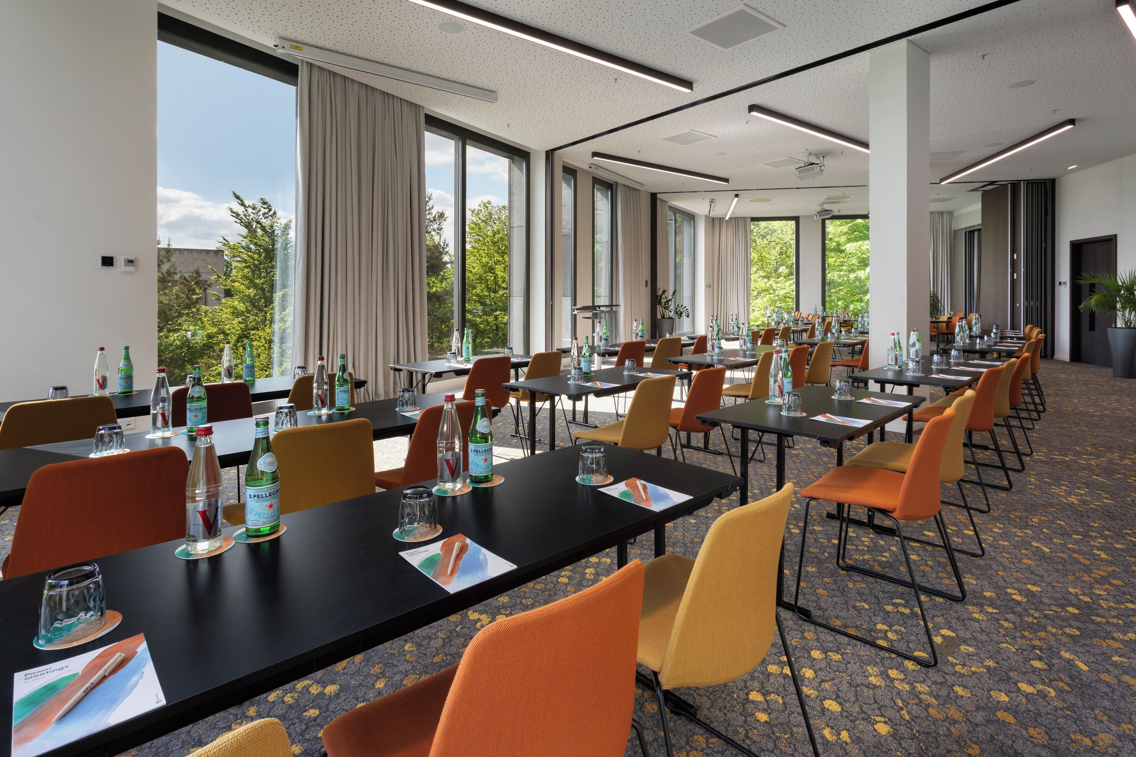 A conference room with multiple tables and chairs, large windows with curtains, and a carpeted floor.