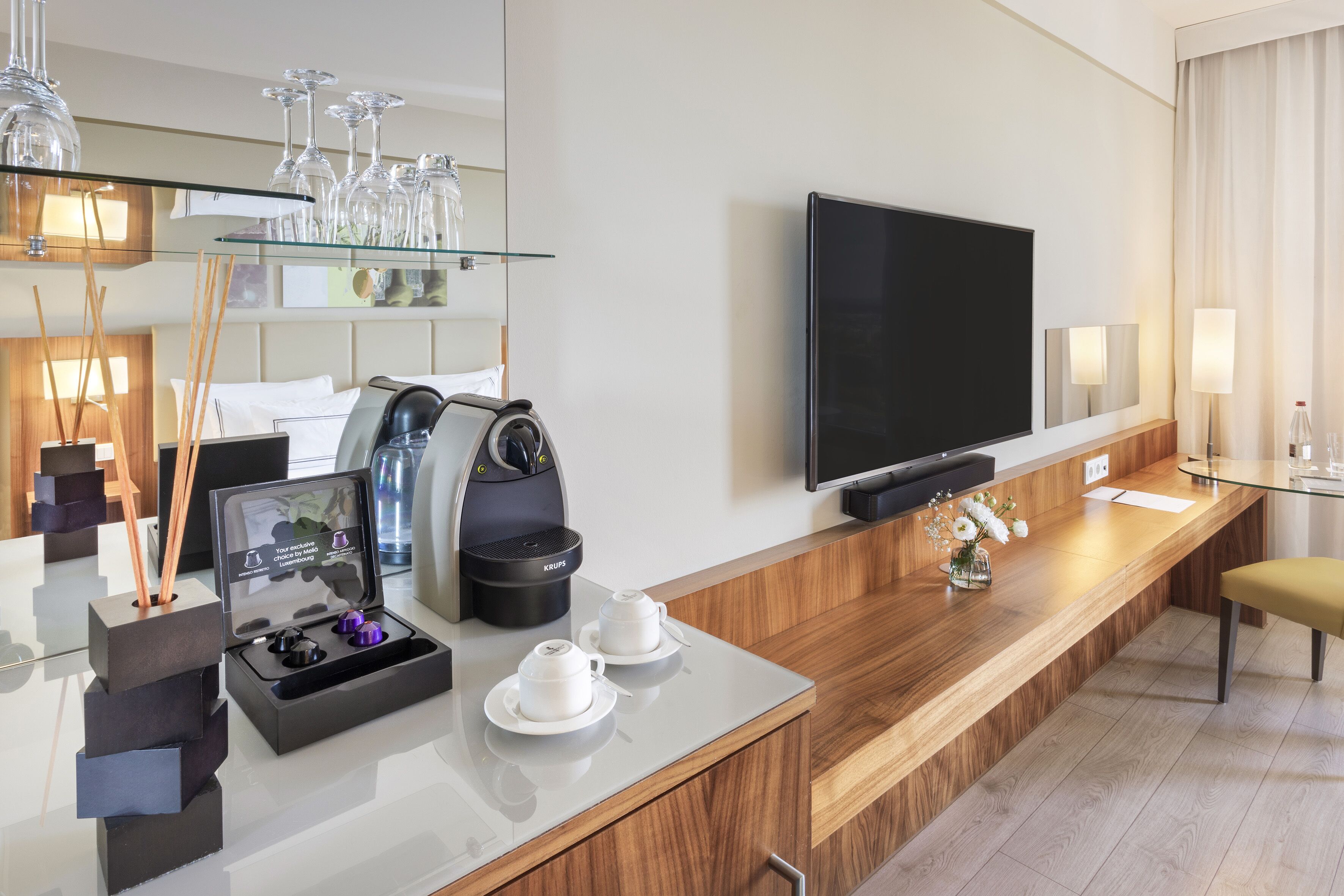 A modern hotel room with a wooden reception desk, a flat-screen TV, a coffee maker, and a glass display case with wine glasses.