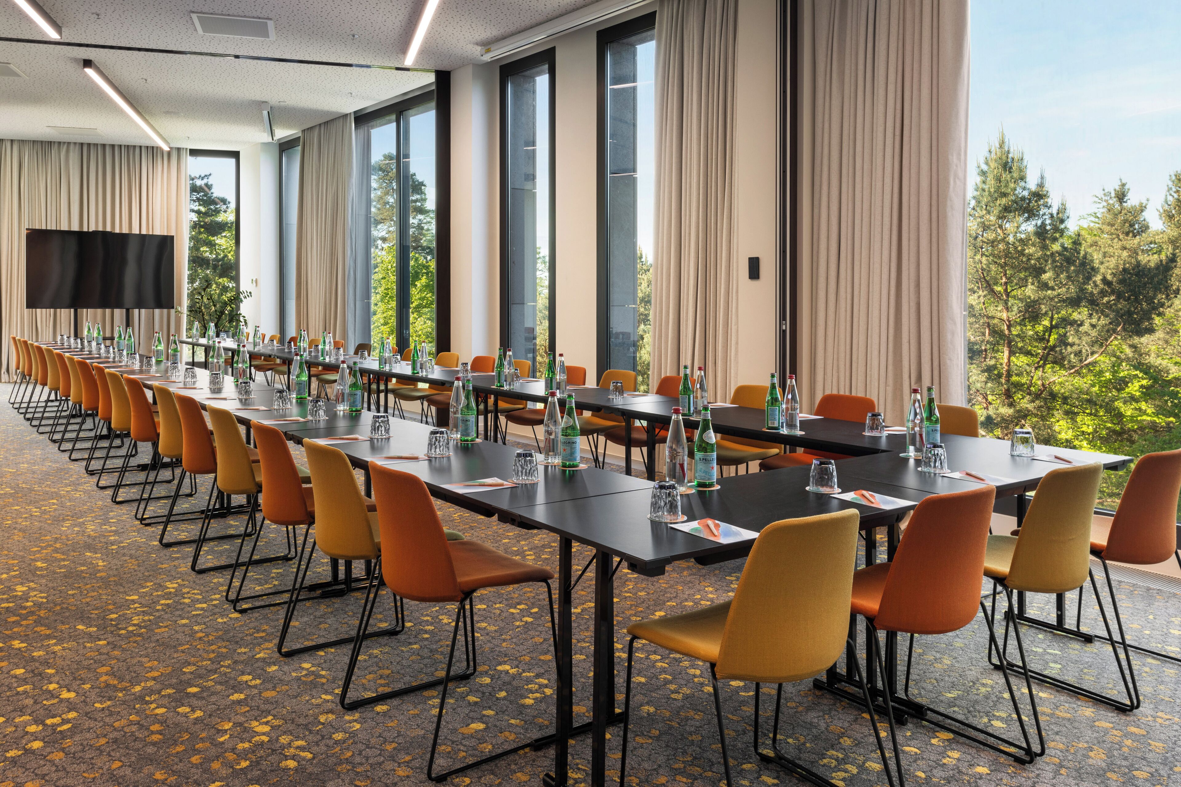 Une salle de conférence avec de longues tables, des chaises orange et de grandes fenêtres dotées de rideaux.