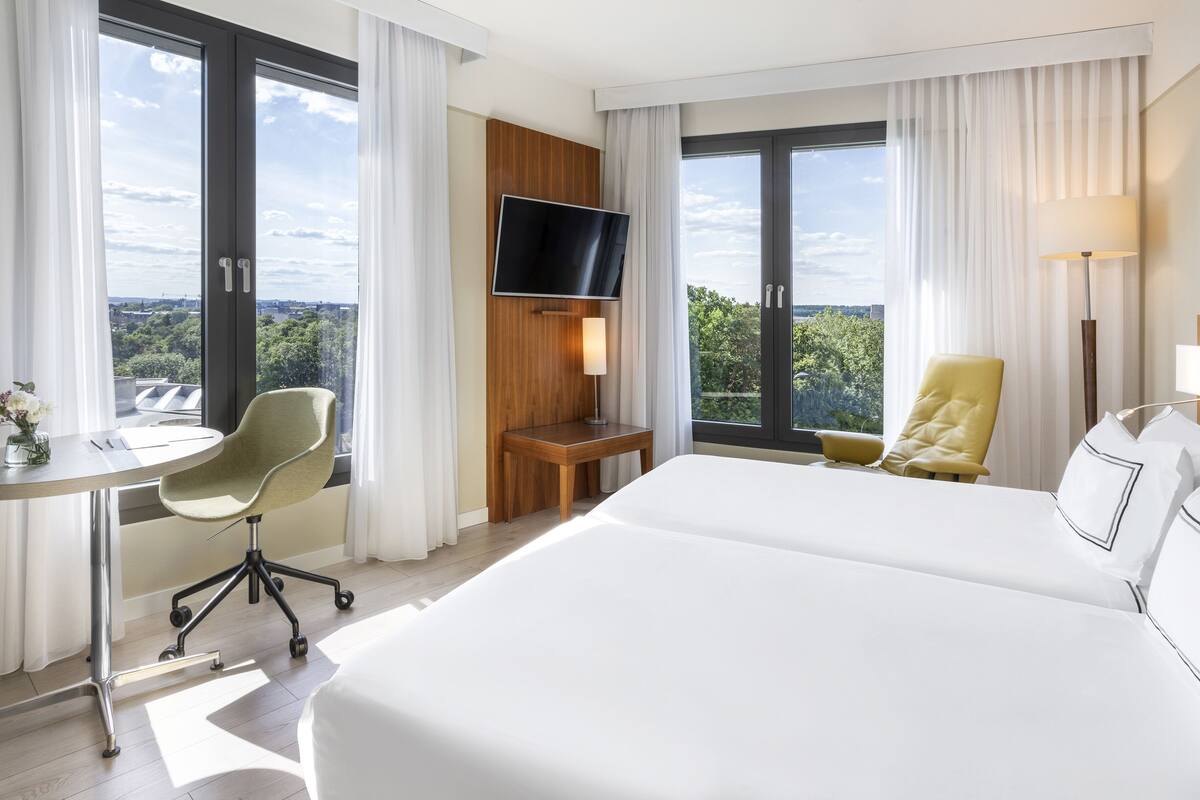 Habitación de hotel con una cama grande, un escritorio con silla, un televisor de pantalla plana en la pared y vistas a la ciudad a través de amplias ventanas.