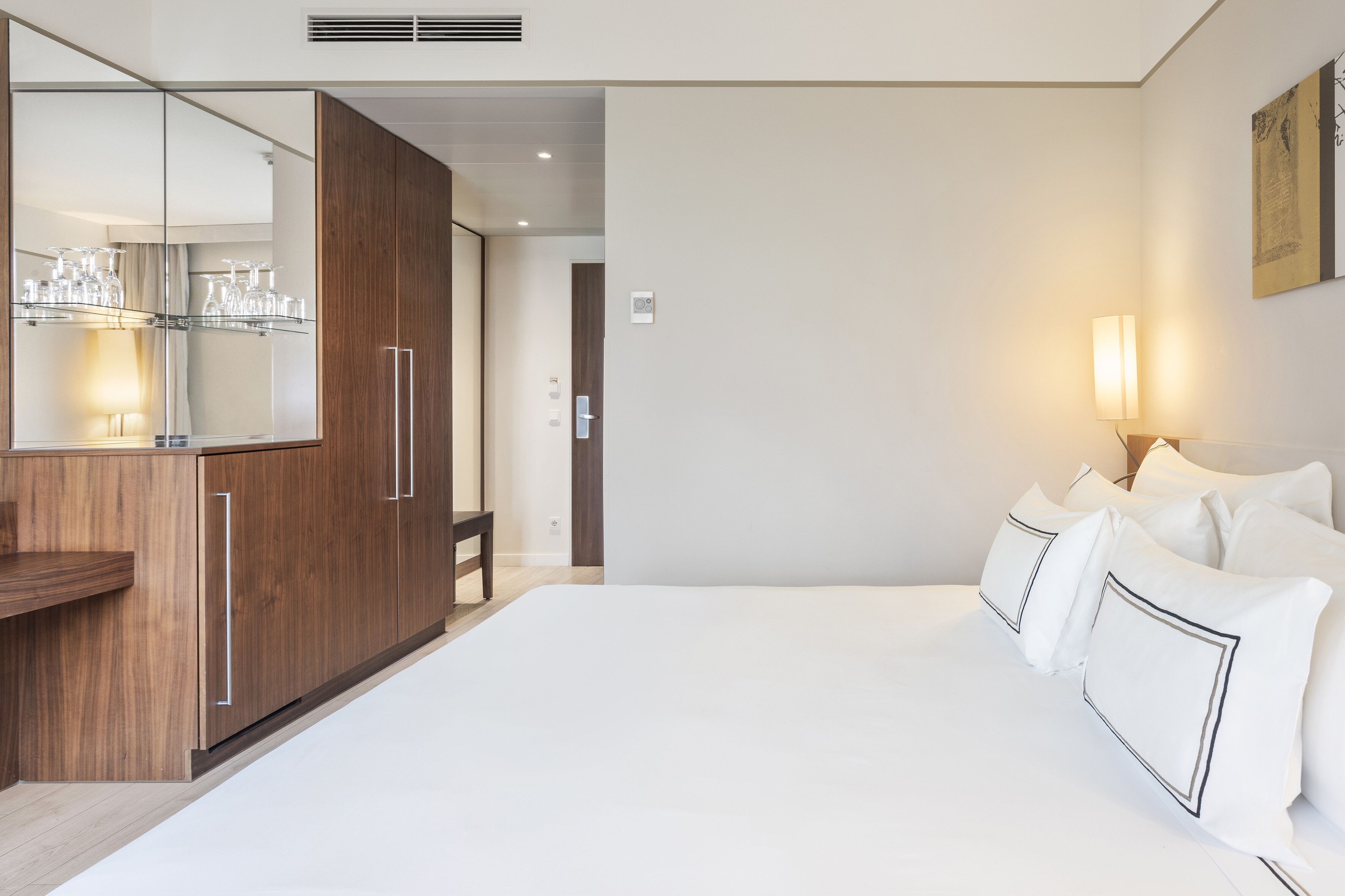 A modern hotel room with a large bed, a wooden wardrobe, a mirror, and a bedside table with glasses.