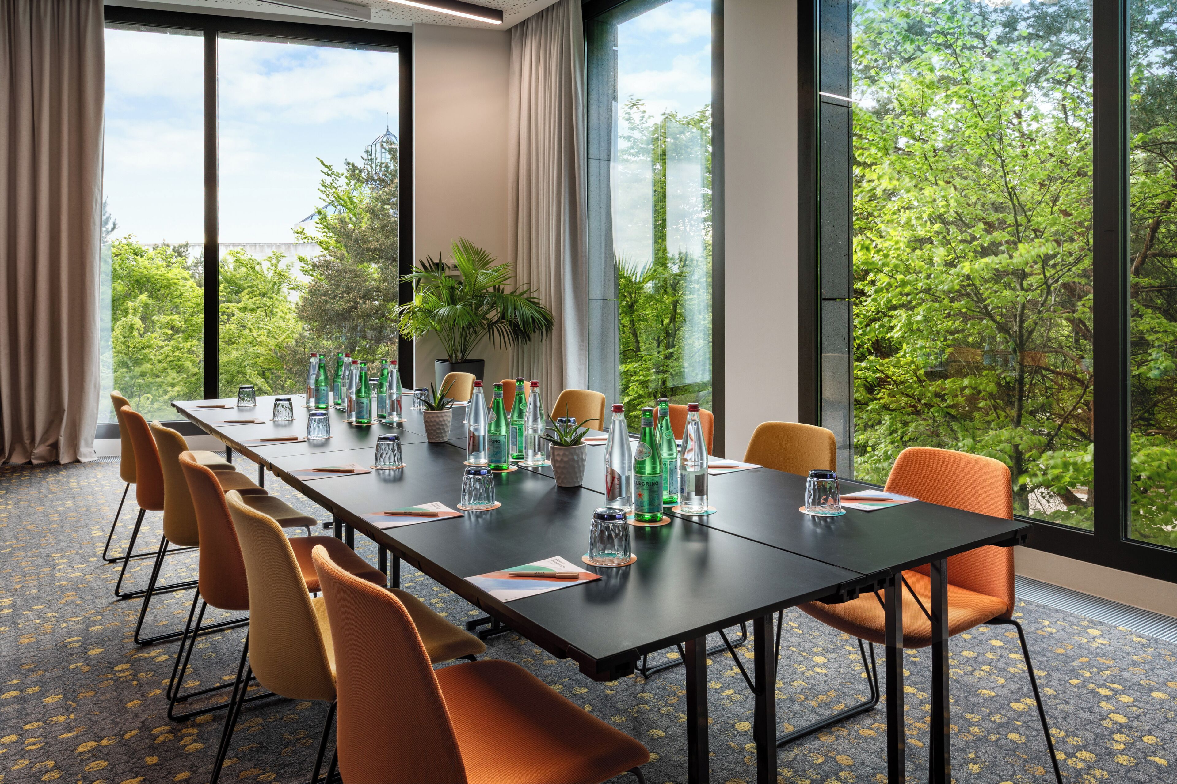 A conference room with a long table, chairs, and a view of greenery through large windows.