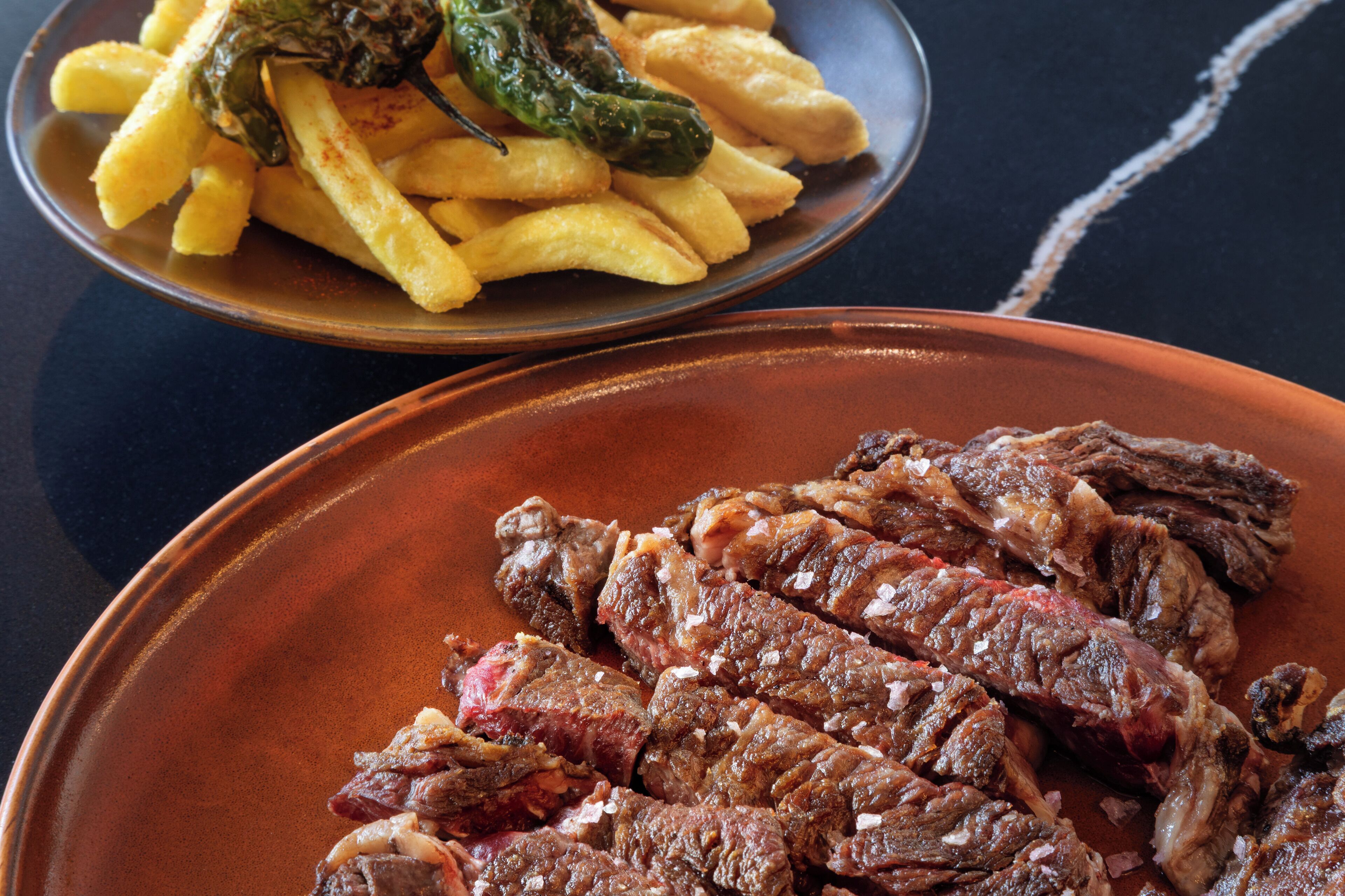 A plate of grilled steak and a bowl of French fries with a green chili.