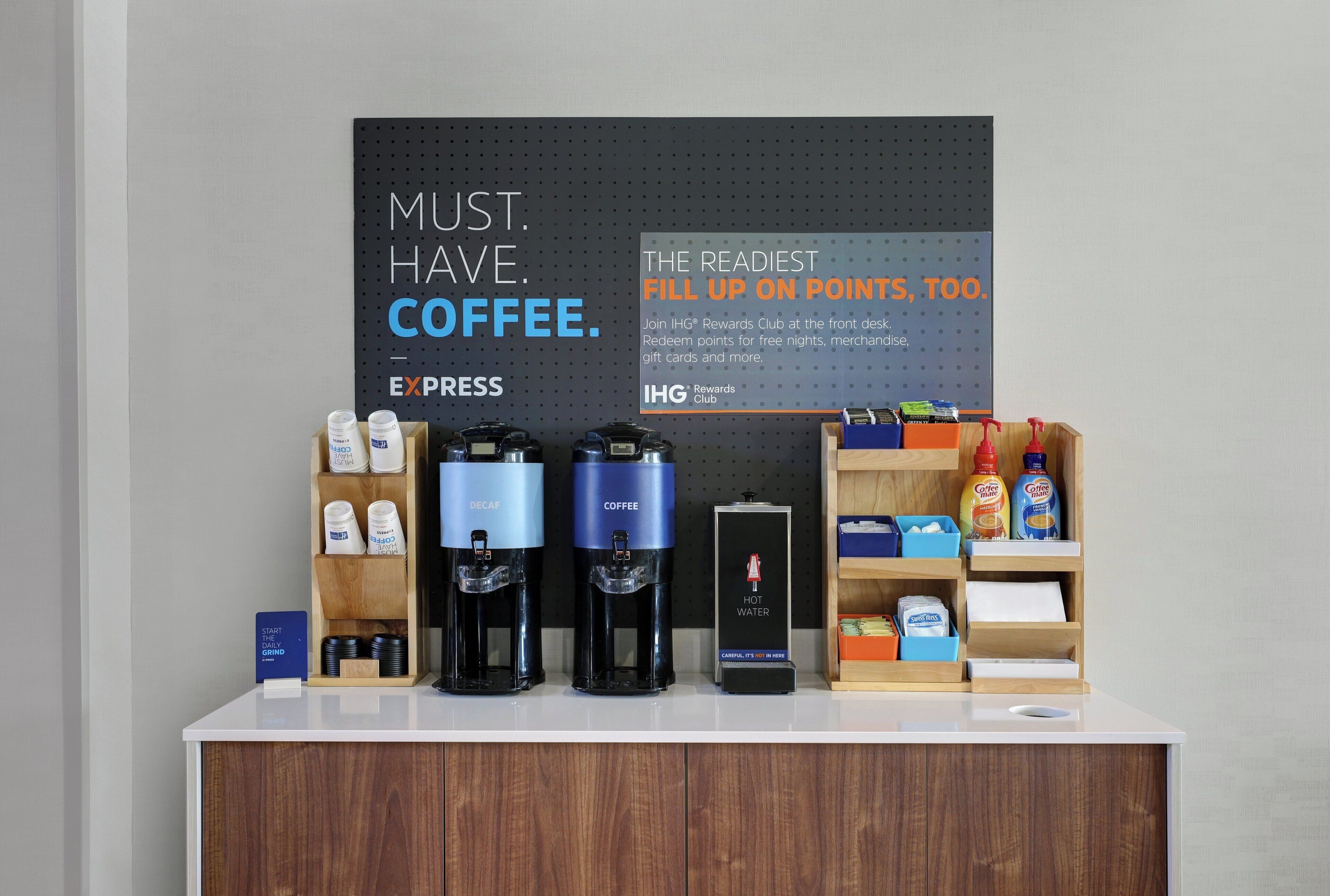 Estação de café com dosadores de marca, máquina de café e placa promovendo um clube de recompensas.