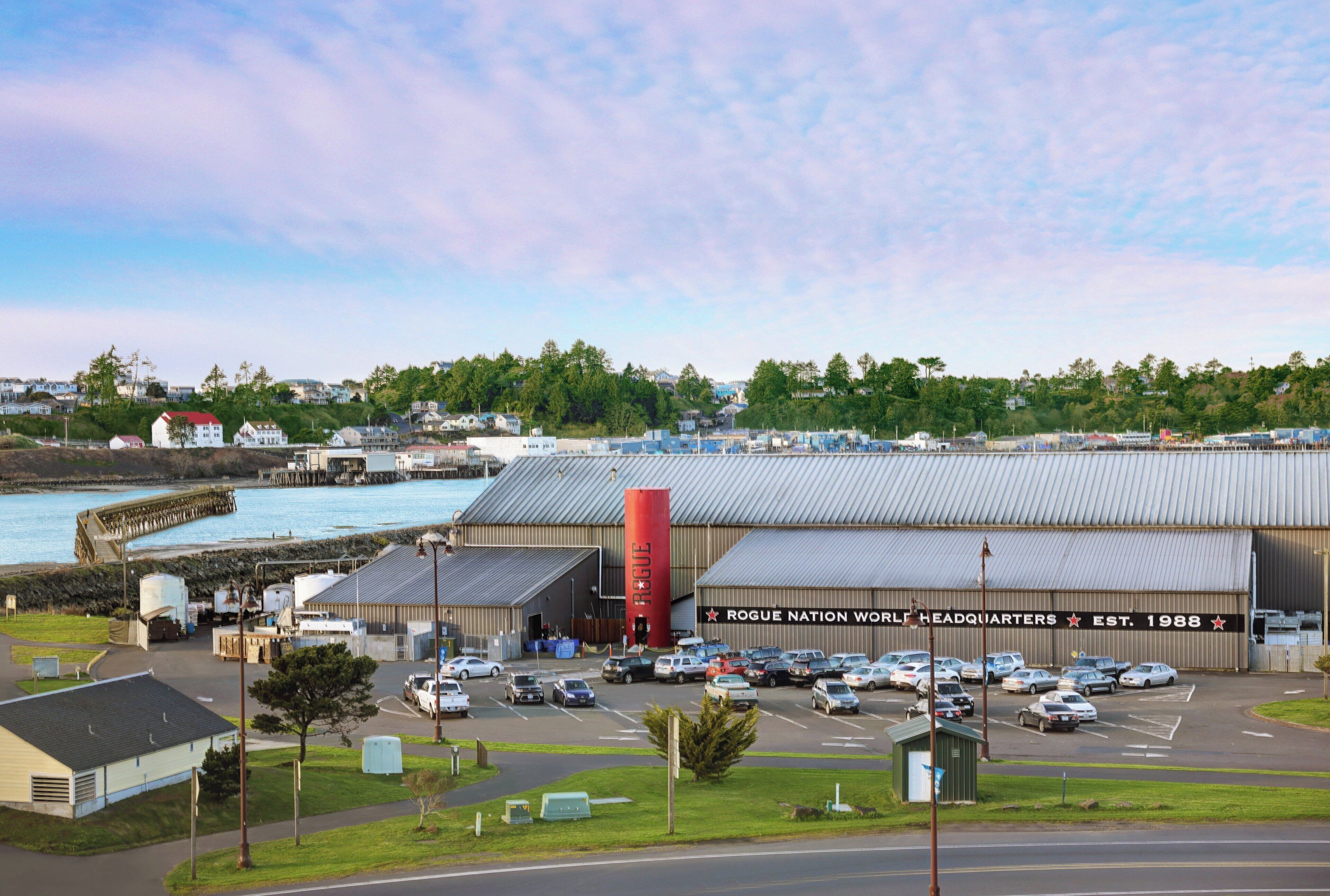 Un grand bâtiment industriel avec un parking à l’avant, une façade donnant sur le port et des immeubles résidentiels en arrière-plan.