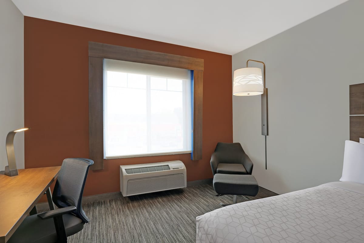A hotel room with a large window, a desk with a chair, a bed, and a standing lamp.