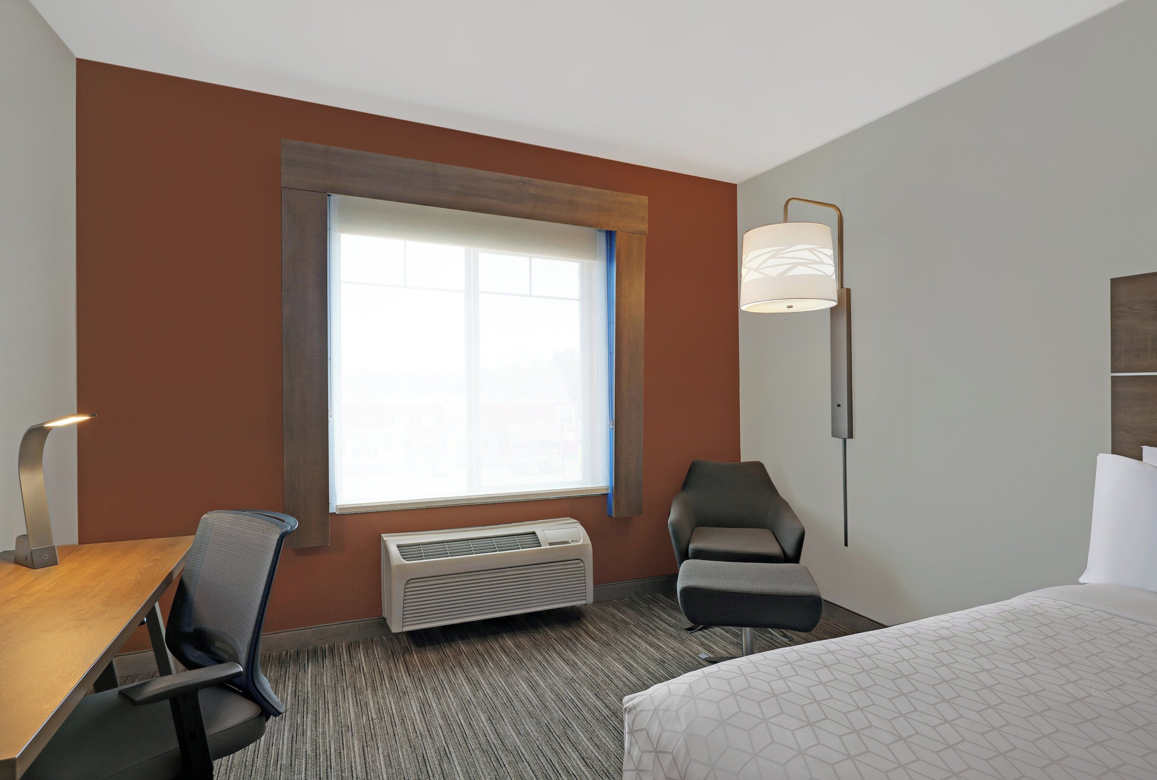 Quarto de hotel com cama, escrivaninha, cadeira, janela, ar condicionado e luminária na parede.