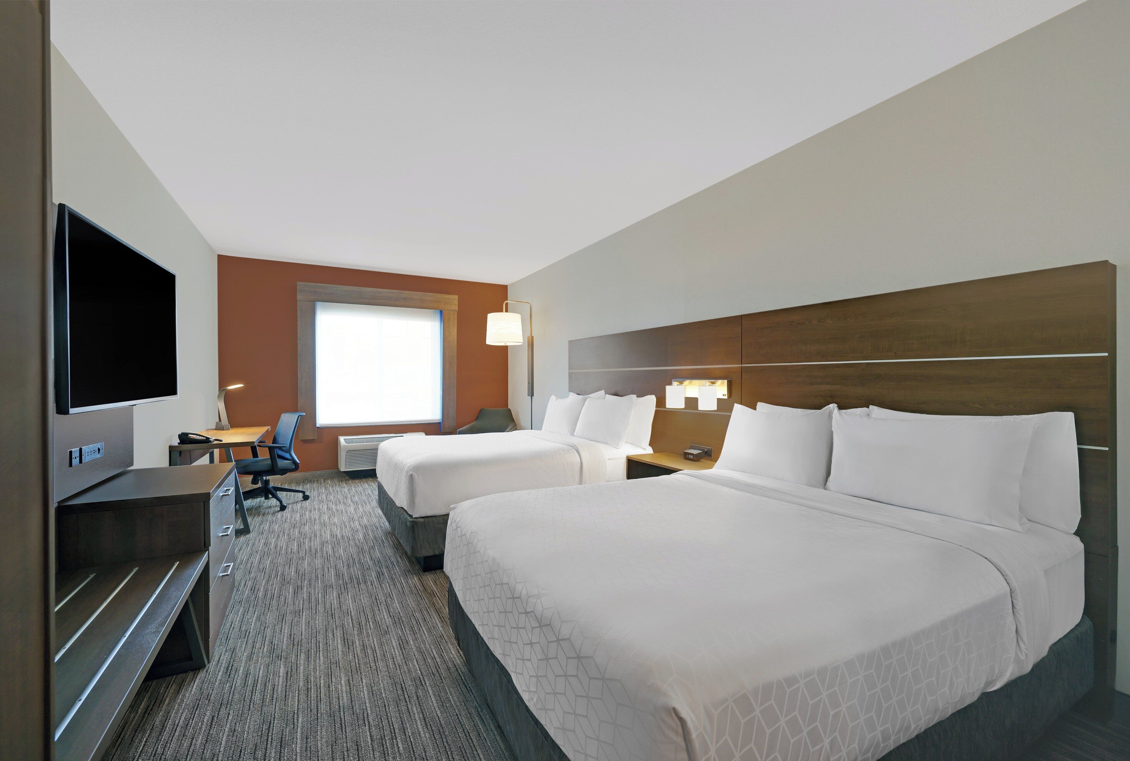 A hotel room with two beds, a desk with a flat-screen TV, a chair, and a window with natural light.