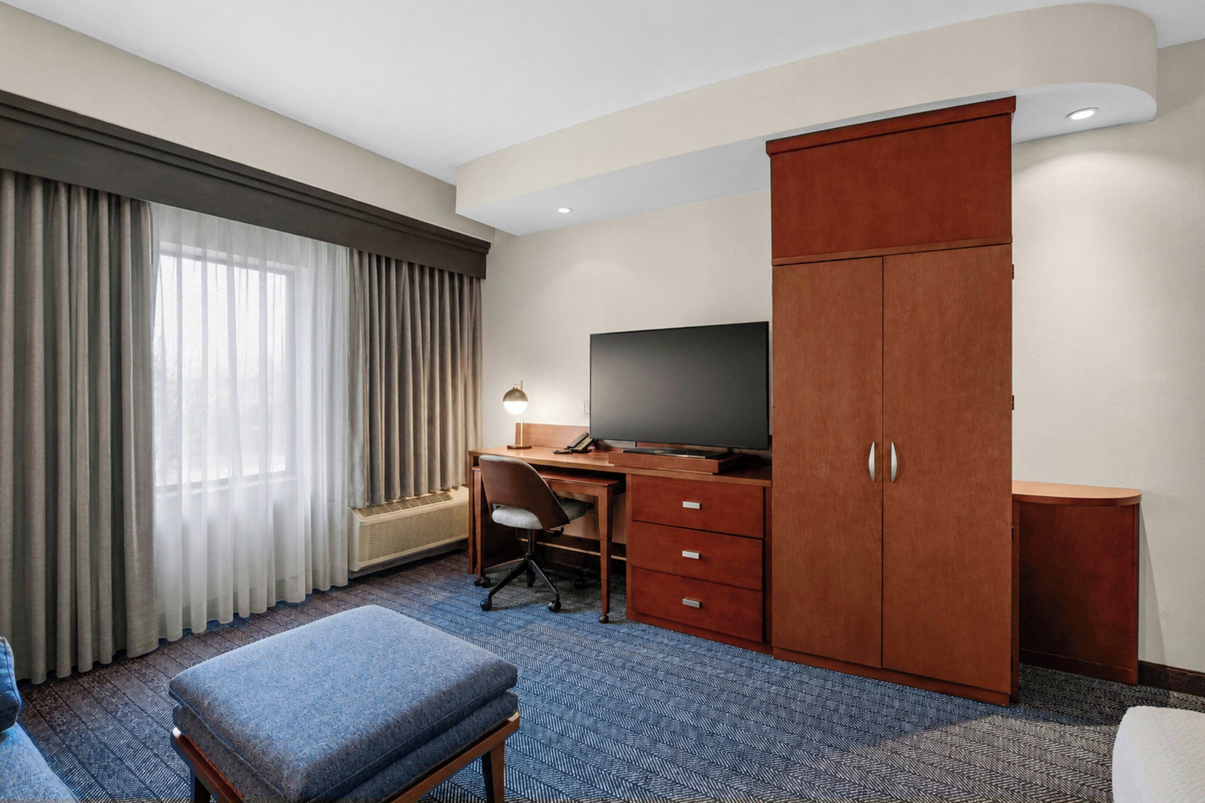 Habitación de hotel con escritorio, televisión, armario y un amplio ventanal con cortinas.