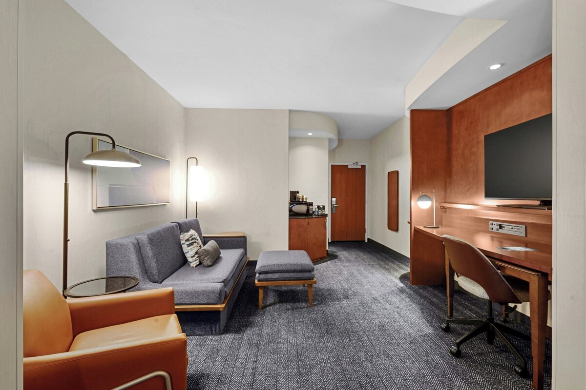 Ein Hotelzimmer mit einer Couch, Hocker, Schreibtisch, Fernseher, Lampe und einer kleinen Küchenzeile.