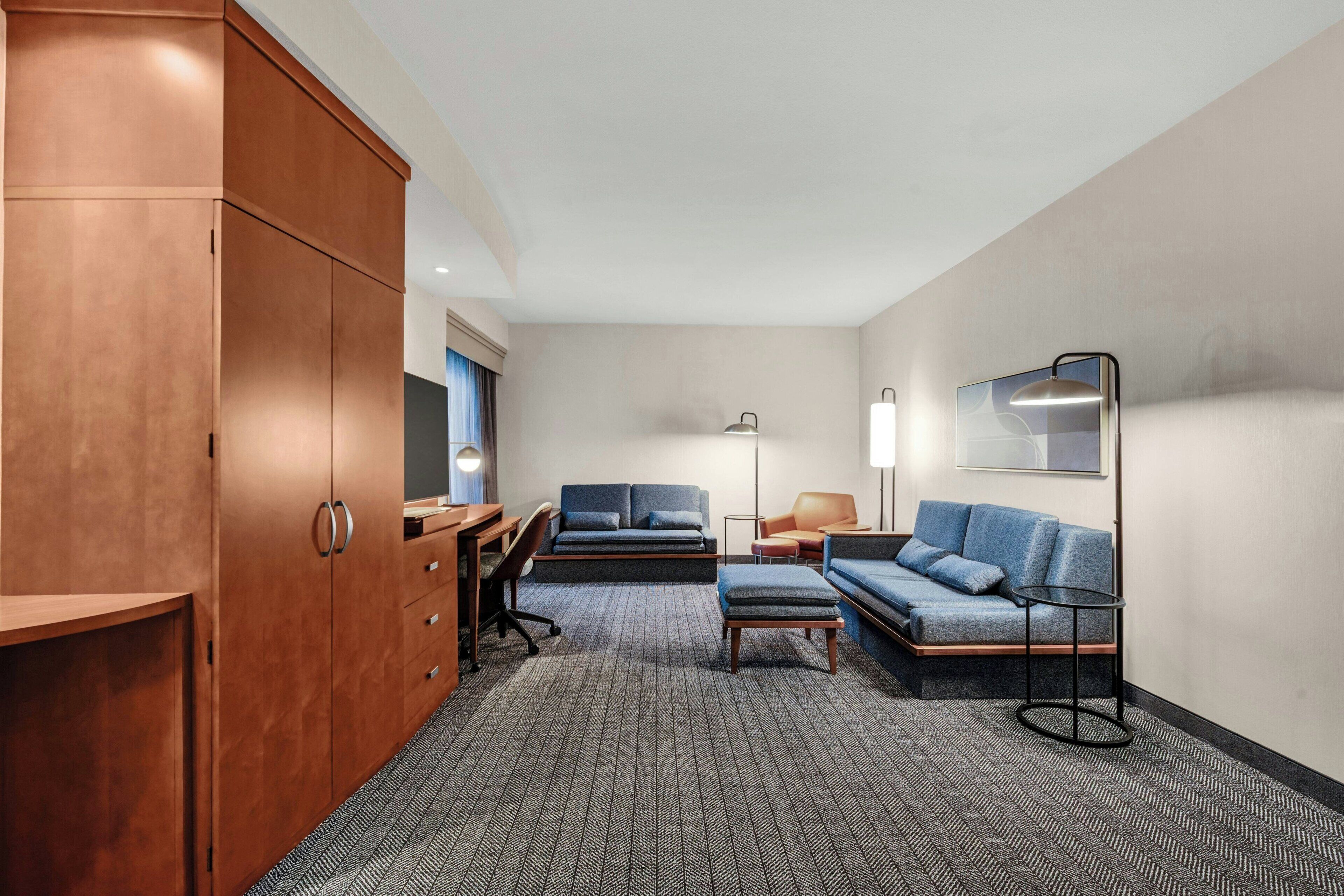 A hotel room with a large wardrobe, a sofa, a desk with a chair, a floor lamp, and a mirror on the wall.