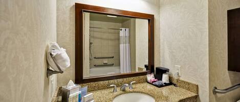 A bathroom with a granite countertop sink, a mirror with a wooden frame, a towel rack, a shower with a glass door, and a coffee maker on the counter.