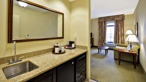 Un baño de hotel con lavamanos, espejo y encimera con artículos de aseo. En la habitación contigua hay una mesita y una silla.