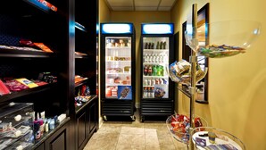 Pasillo de hotel con dos refrigeradores iluminados llenos de bebidas, un bar de aperitivos con diversos productos envaselados y una exhibición de artículos de tocador.