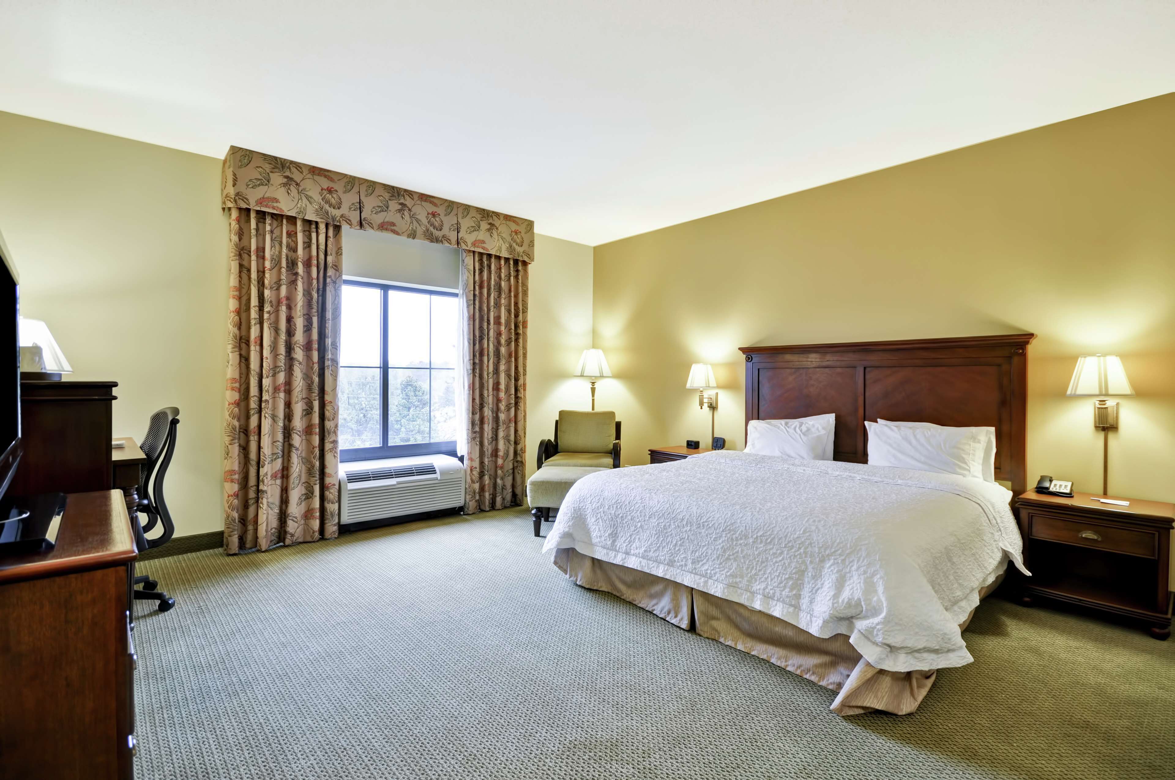 Habitación de hotel con una cama grande, dos lámparas de noche, un escritorio con una silla, una ventana con cortinas y un equipo de aire acondicionado.