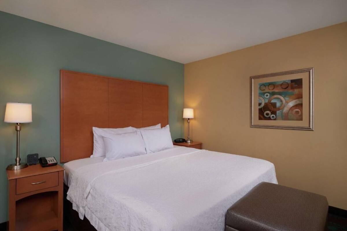 A hotel room with a bed, bedside lamps, a nightstand, a telephone, and a framed abstract painting on the wall.