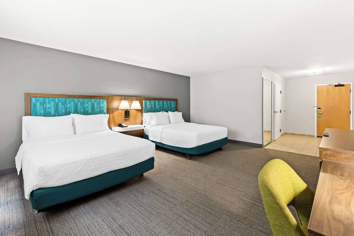 A hotel room with two beds, a desk, a chair, and a door.