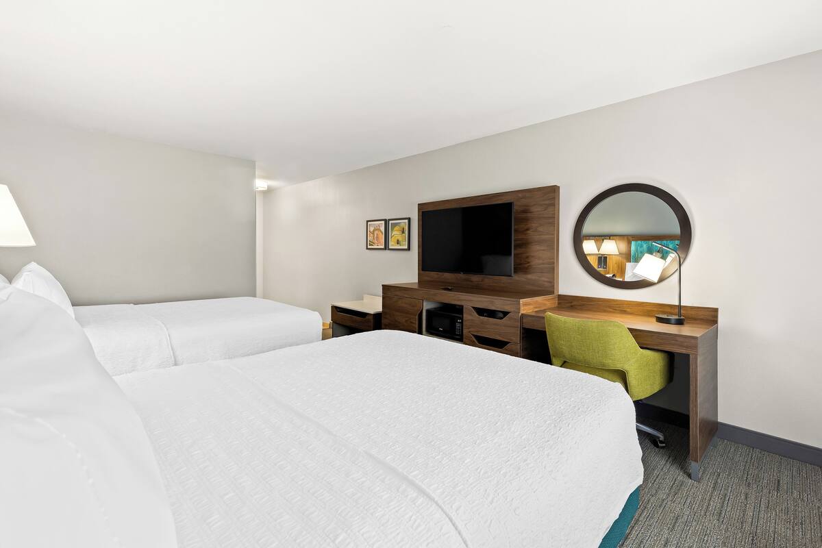 A hotel room with two beds, a desk, a flat-screen TV, a mirror, and a lamp.