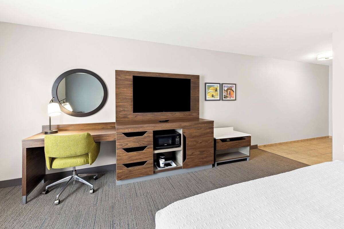 A hotel room with a wooden desk, a green chair, a flat-screen TV, a microwave, a small shelf, and a bed.