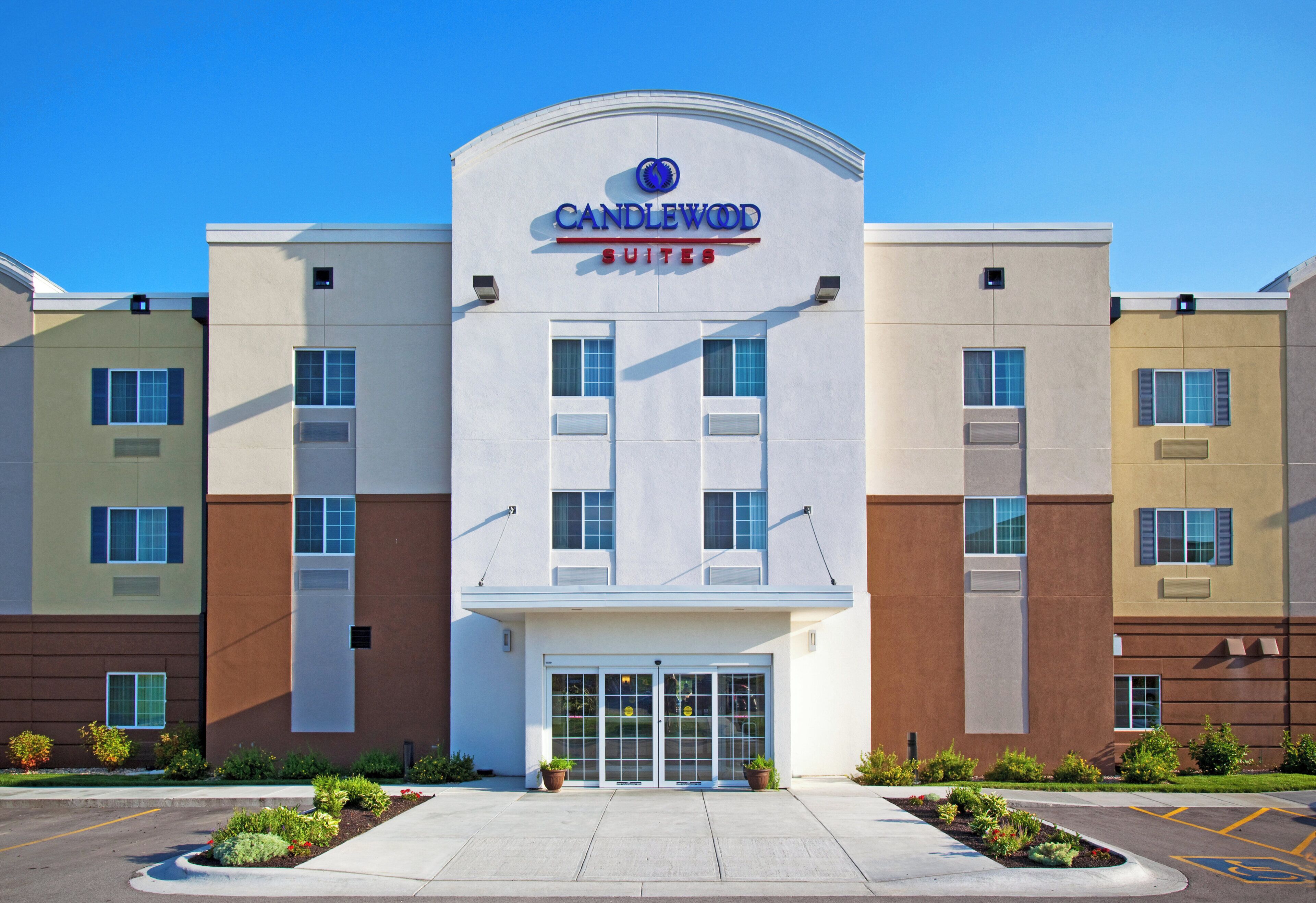 Un hôtel à plusieurs étages, reconnaissable à son grand panneau « CANDLEWOOD SUITES », doté d’une entrée principale avec une porte en verre et d’un parking avec des places délimitées.