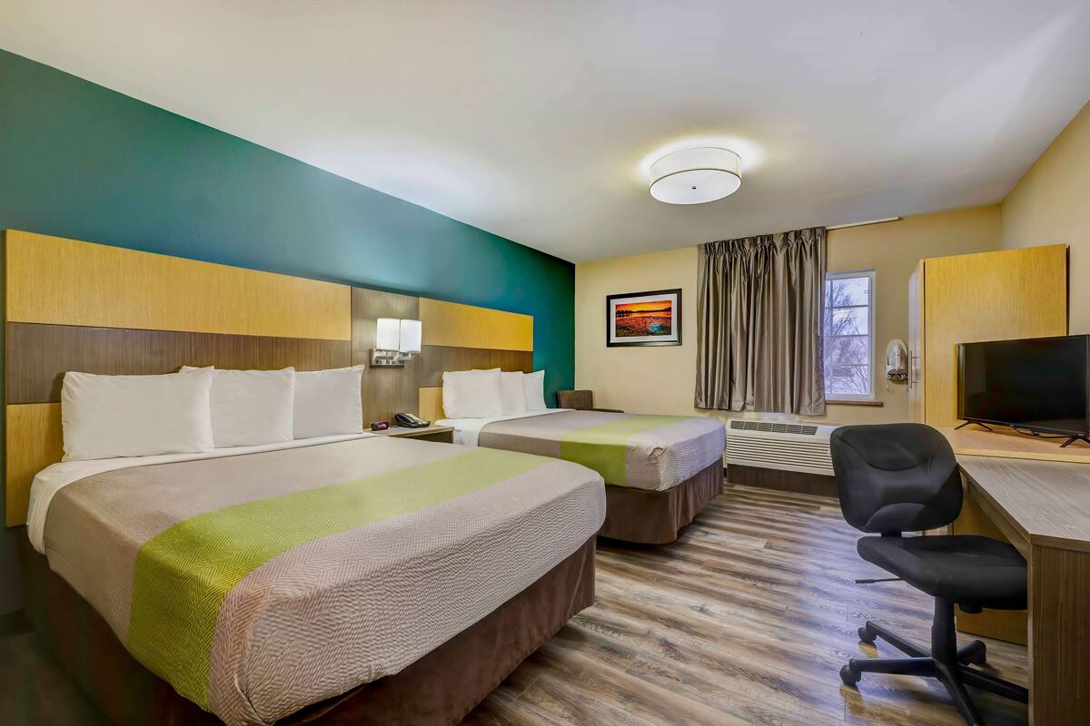 A hotel room with two beds, a desk with a chair, a television, a window with curtains, and a painting on the wall.