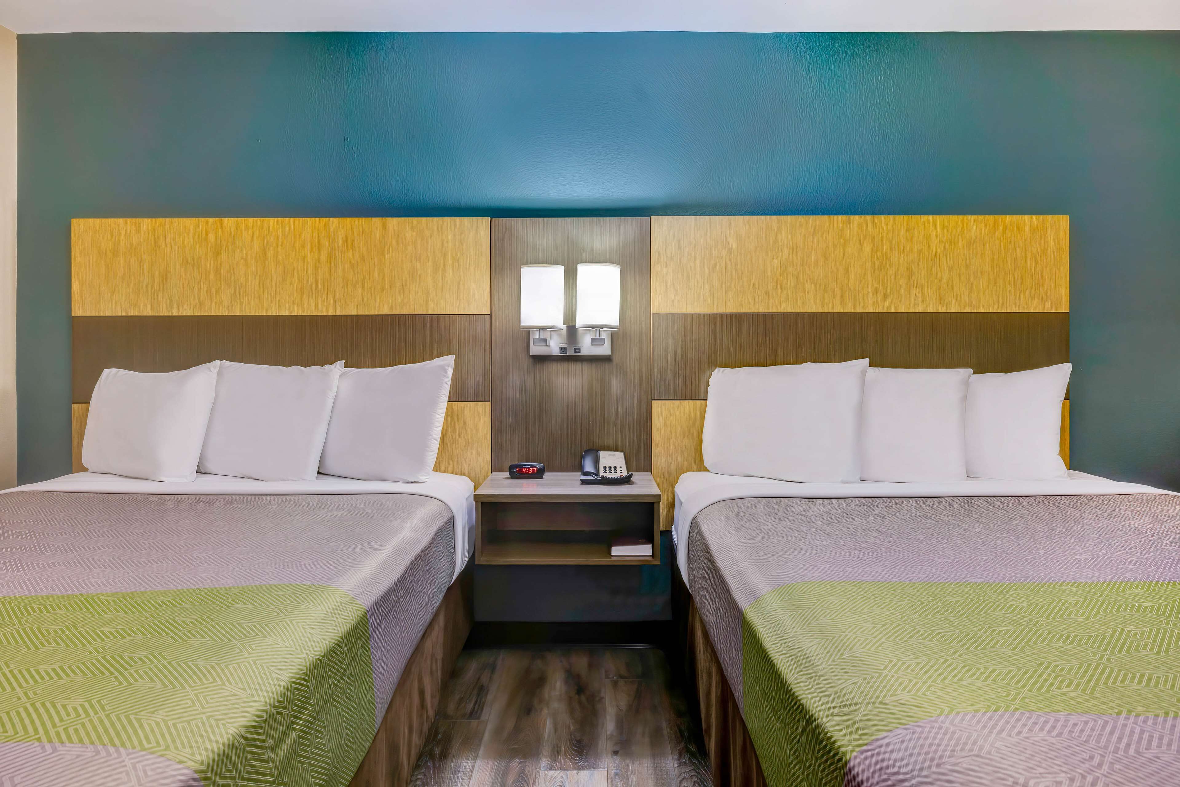 Habitación de hotel con dos camas, una mesita de noche con teléfono y reloj, y una lámpara de pared.