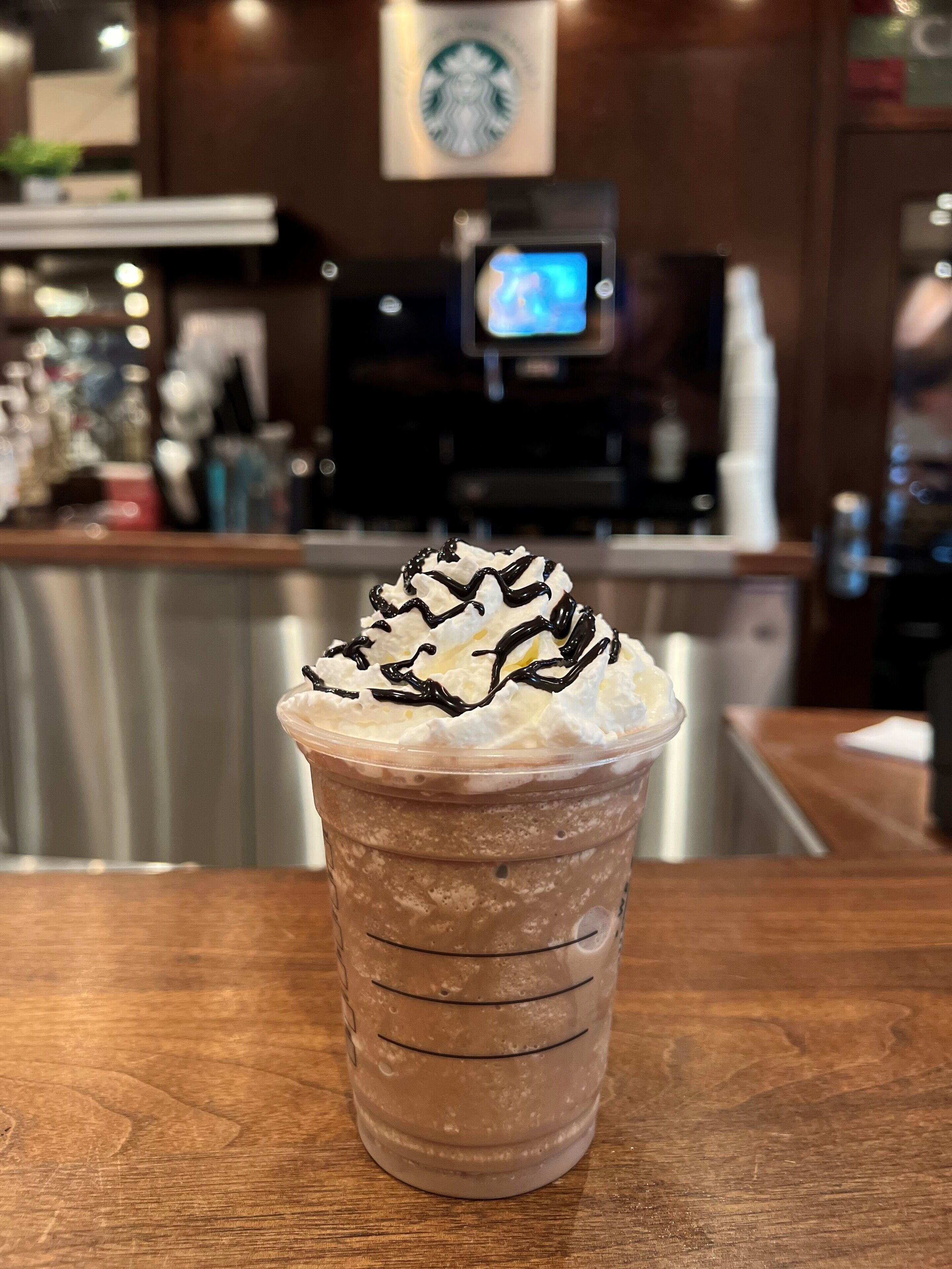 Una bebida en vaso de plástico transparente con crema batida y un chorrito de chocolate sobre una mesa de madera.