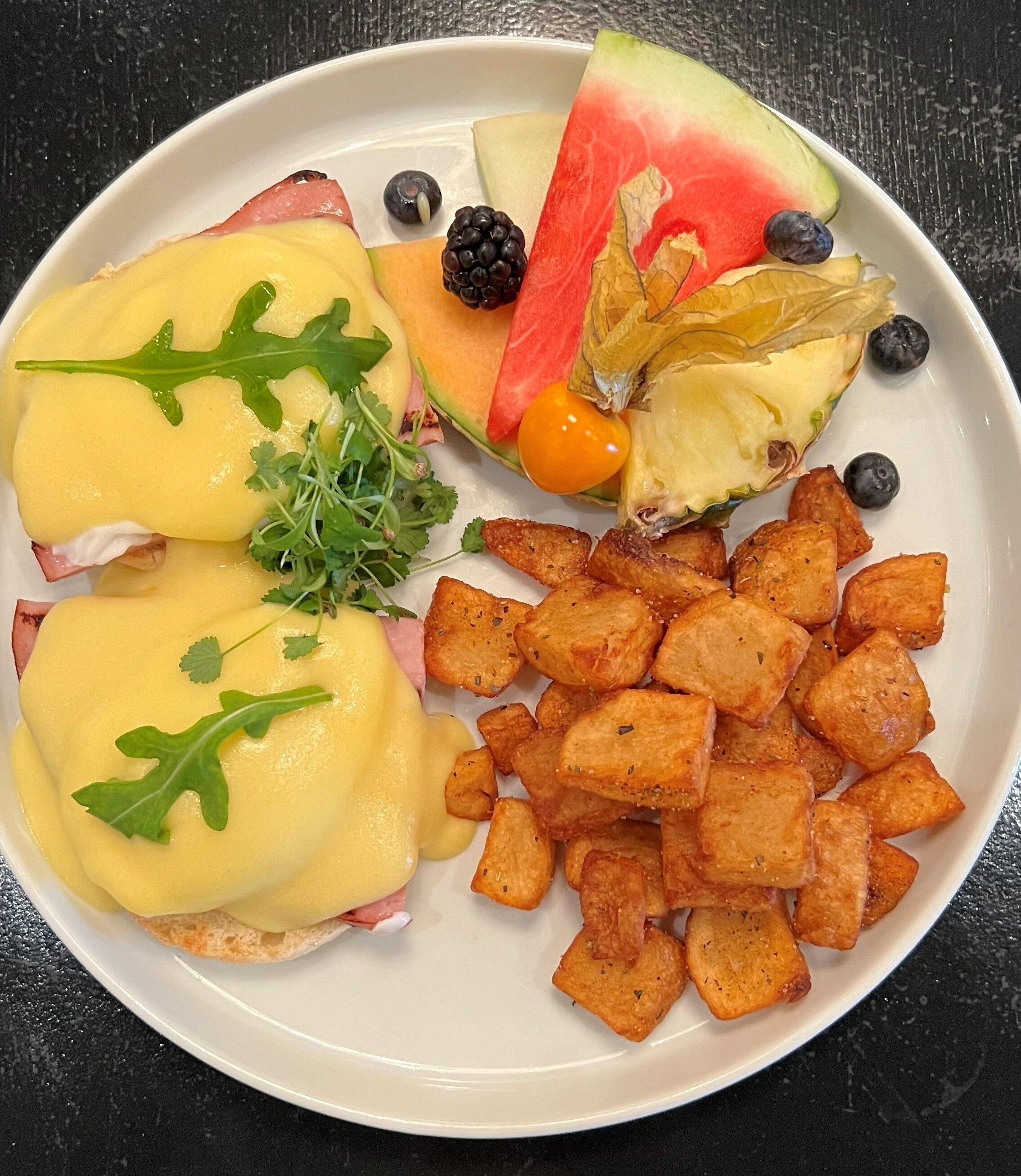 A plate with eggs benedict, roasted potatoes, and fresh fruit.
