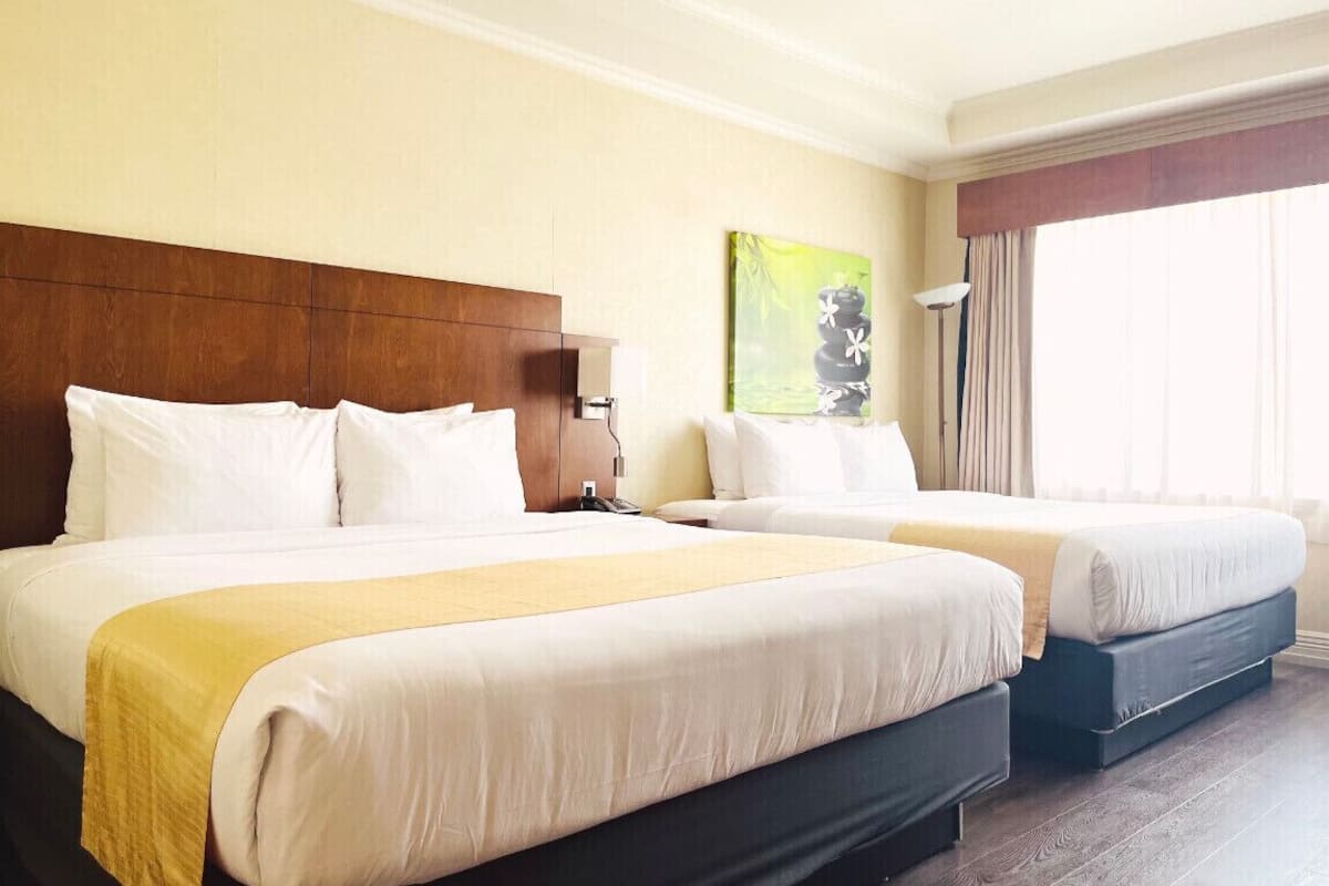 Une chambre d’hôtel avec deux lits, une tête de lit en bois, un tableau au mur et une fenêtre avec des rideaux.