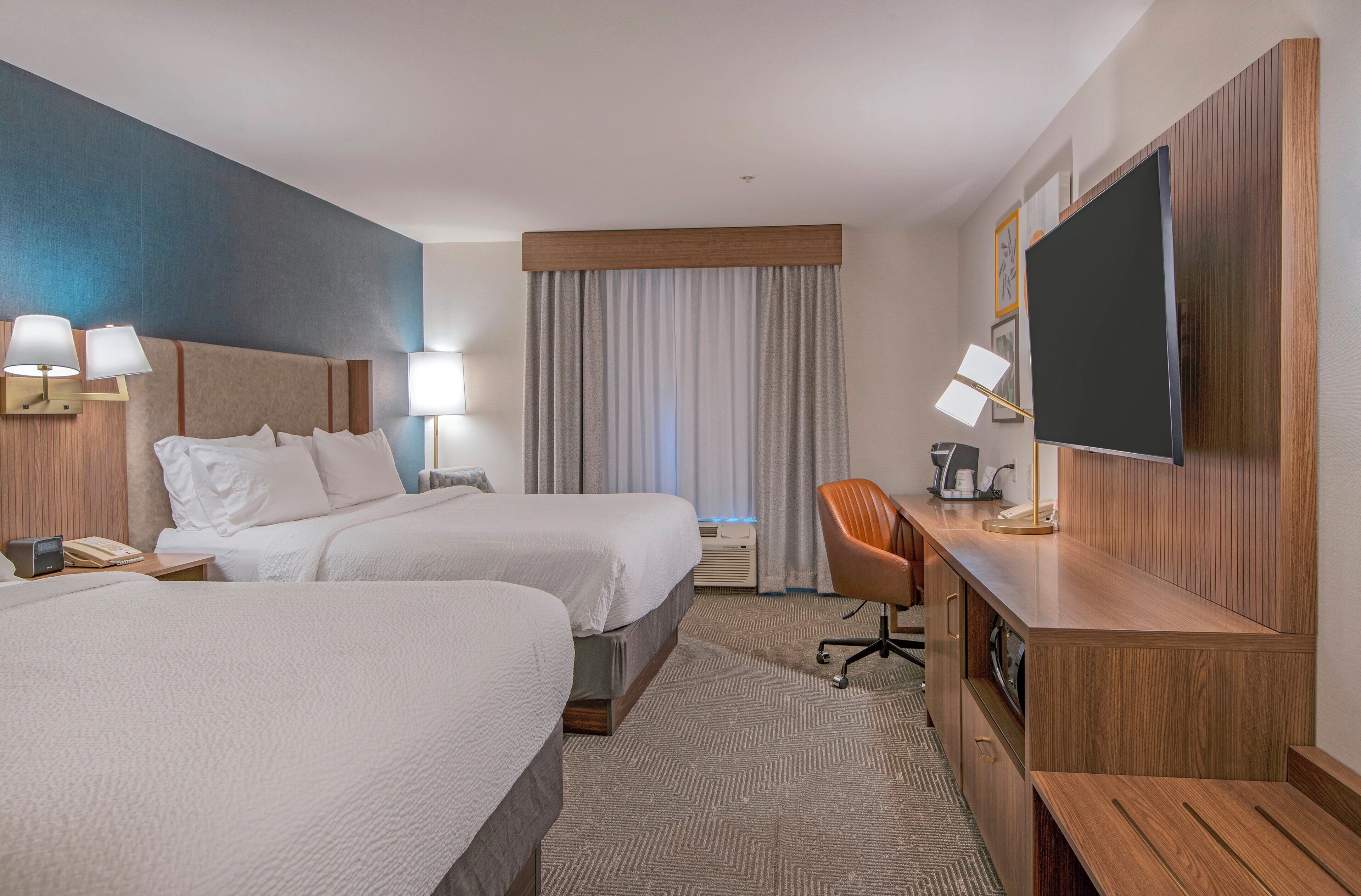 A hotel room with two beds, a desk, a TV, and a large window with curtains.
