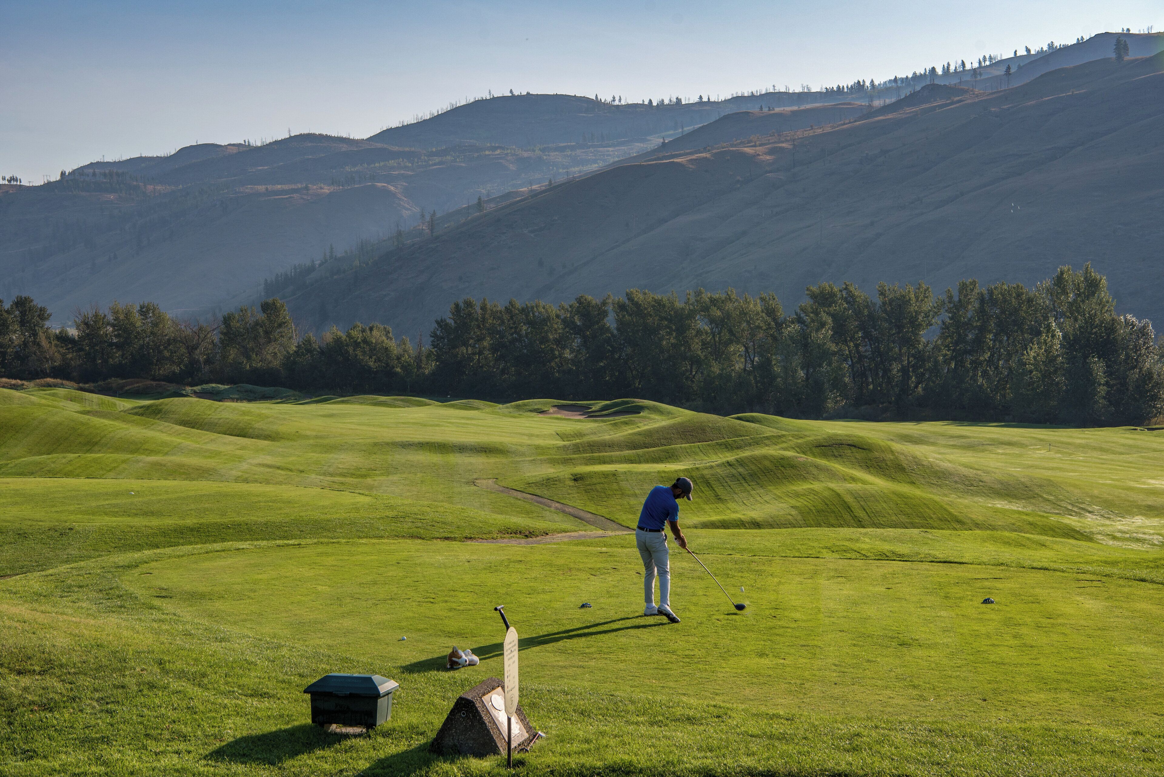A golf course with rolling green hills, a golfer in mid-swing, and a flagstick in the foreground.