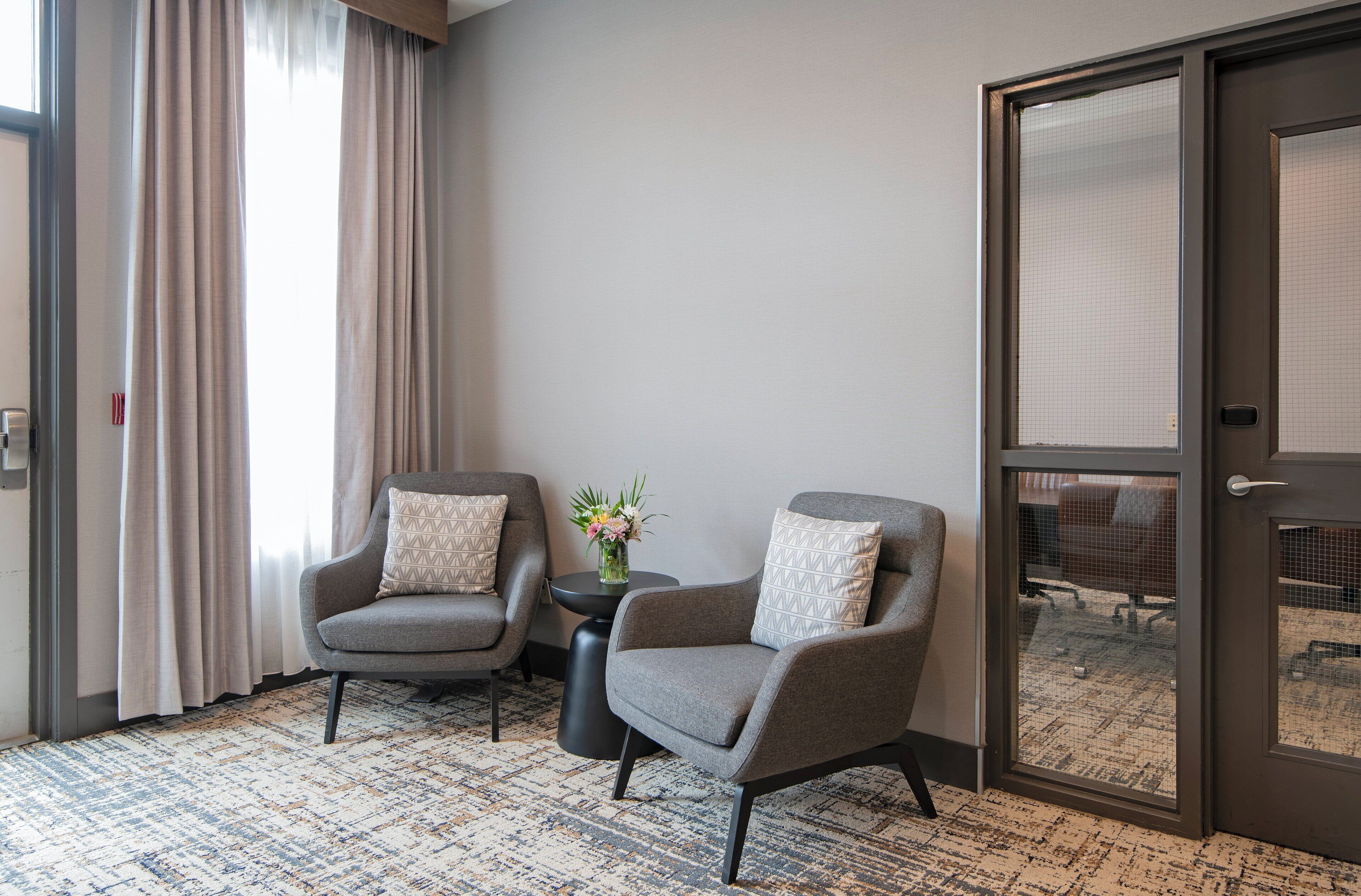 A hotel room with two grey armchairs, a small round table with a vase of flowers, a patterned carpet, and a large window with curtains.