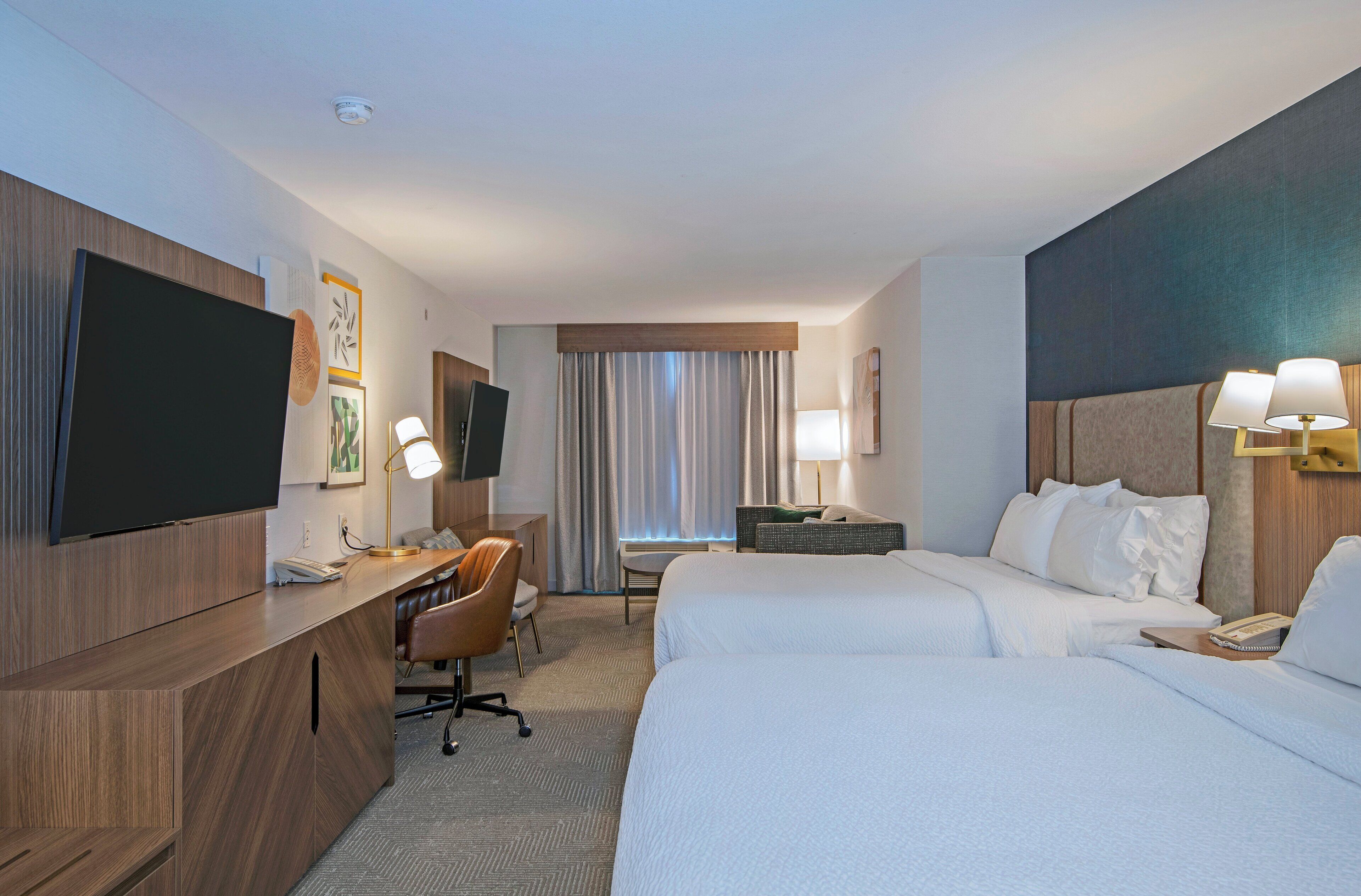 A hotel room with a bed, a desk with a chair, a television, and a window with curtains.