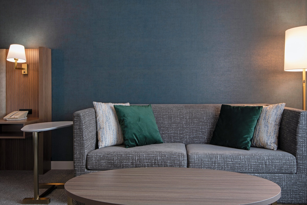 A modern hotel room with a grey textured sofa, two green pillows, a small round table, and two wall-mounted lamps.