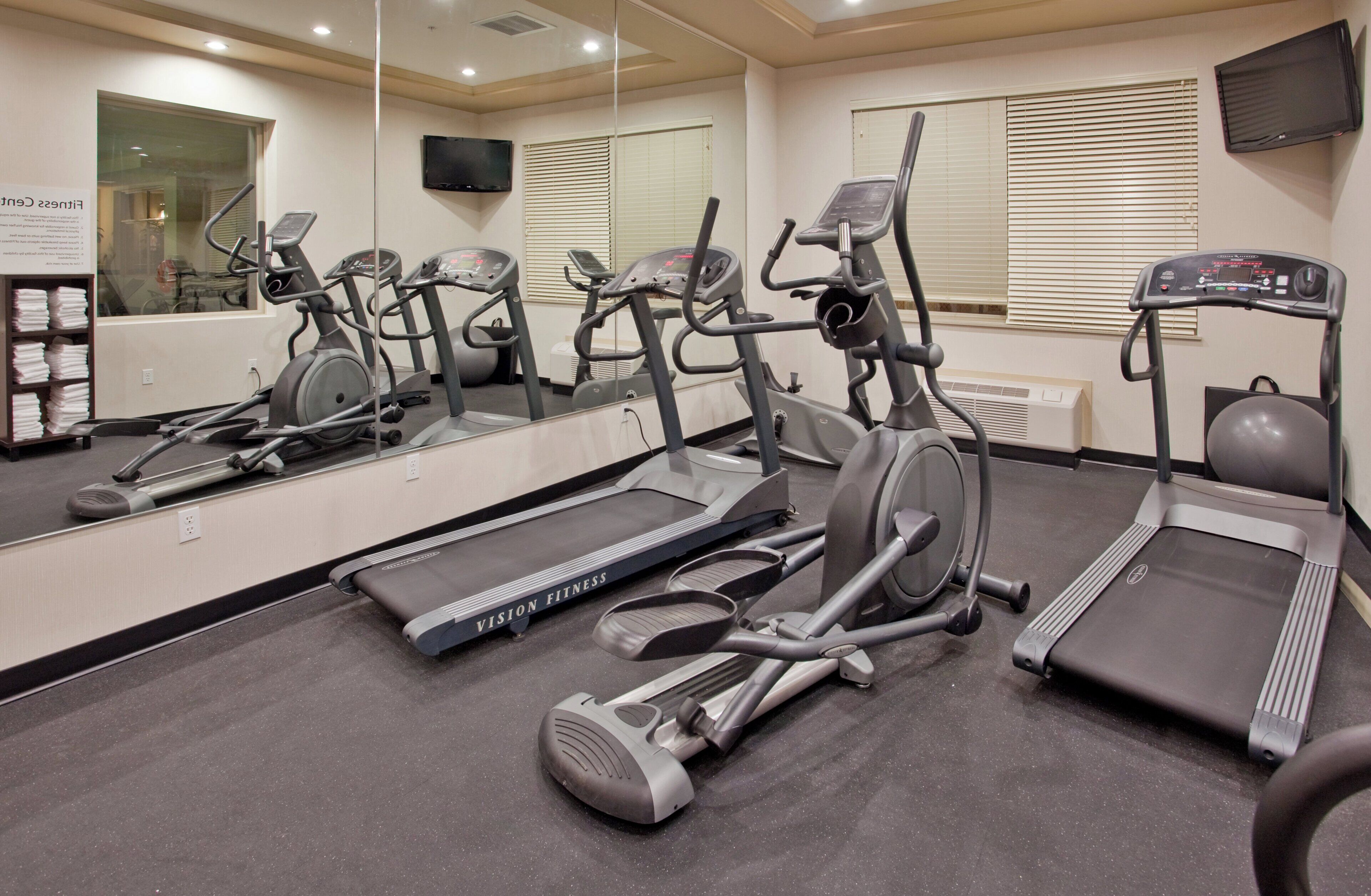 A gym with multiple exercise machines, including treadmills and elliptical trainers, and a large mirror reflecting the equipment.