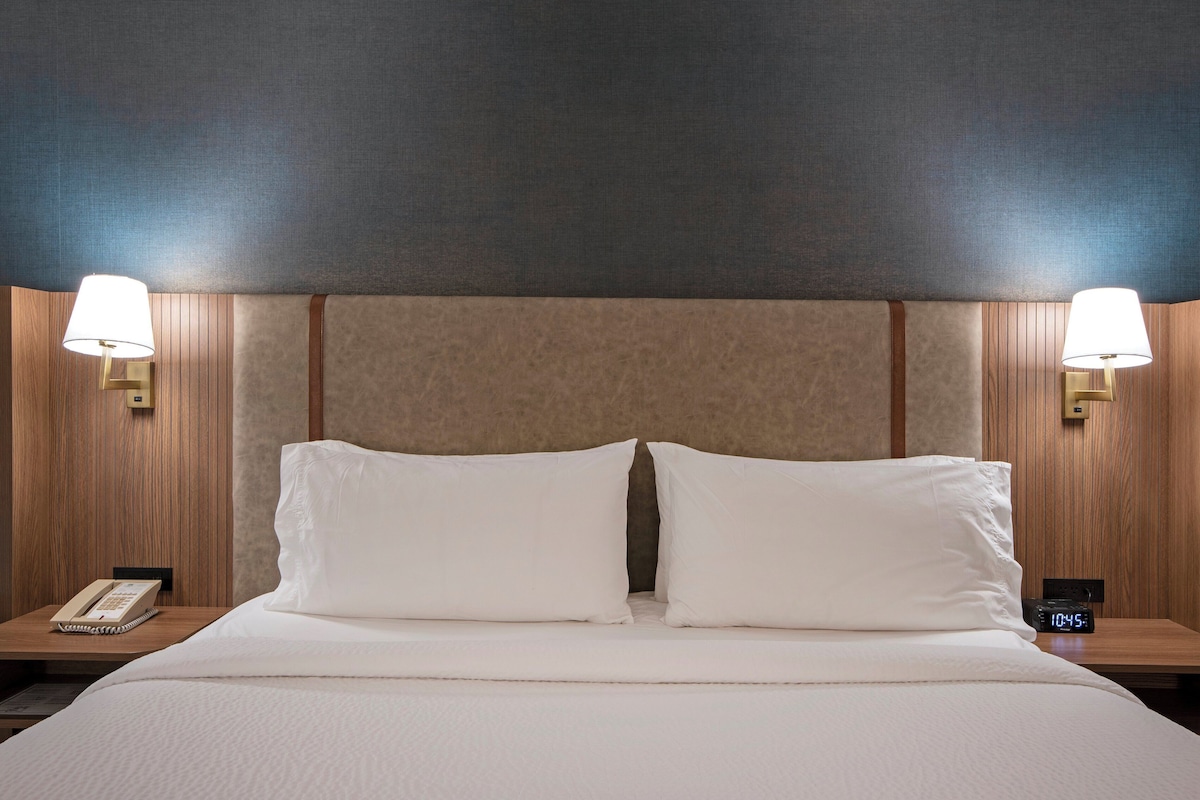 A hotel room with a large bed, two bedside lamps, a nightstand with a phone, and a digital clock.