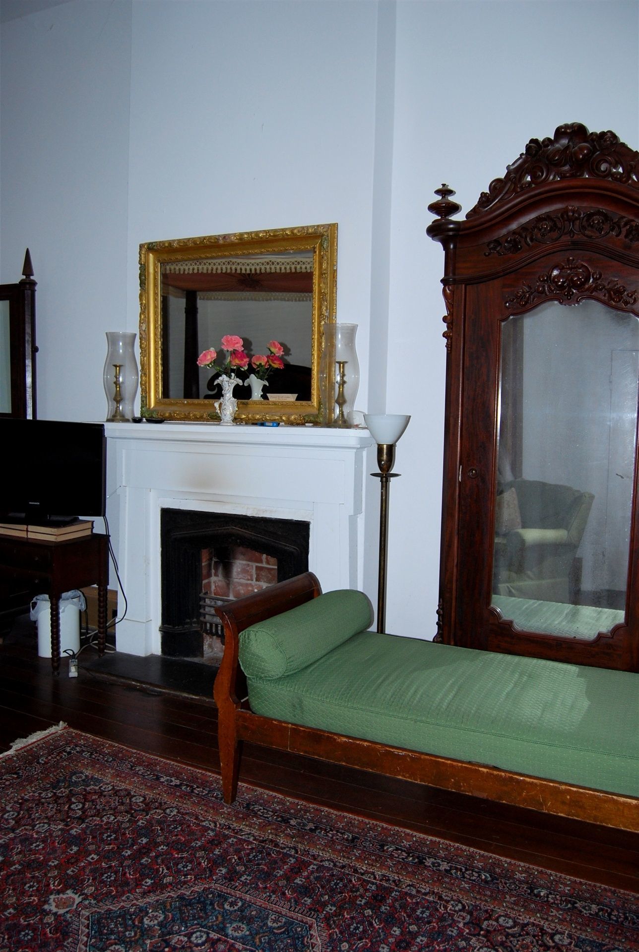 A room with a green upholstered bed, a large mirror with an ornate frame, a fireplace with a white mantel, and a television on a stand.