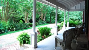 A covered porch with wicker furniture, surrounded by lush greenery and a brick pathway.