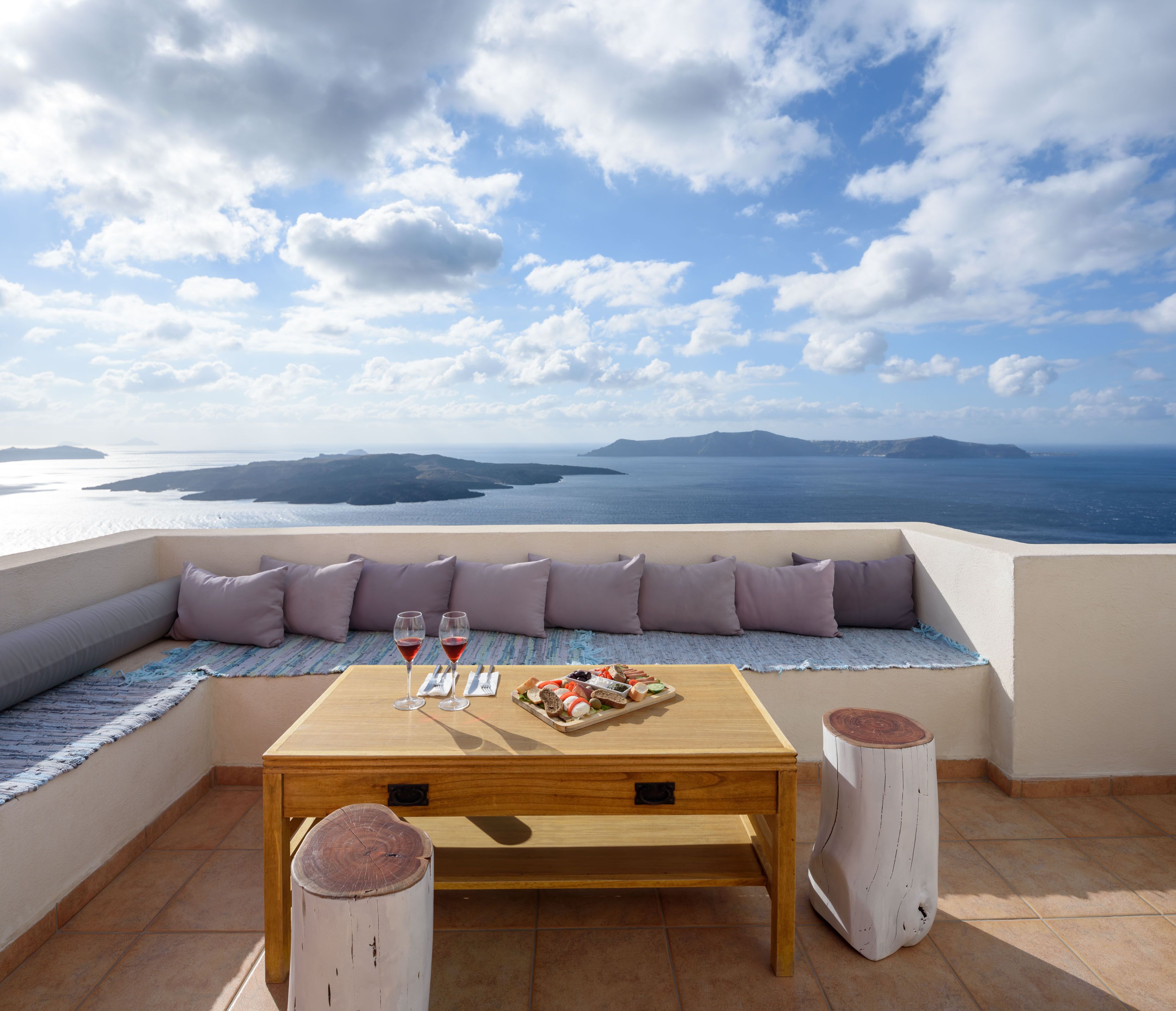 A terrace with a wooden table, two glasses of red wine, a tray of food, and a view of the sea and islands.