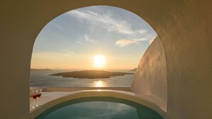 Una ventana circular con vistas a un atardecer sobre un cuerpo de agua, con dos copas de vino tinto en un repisa.