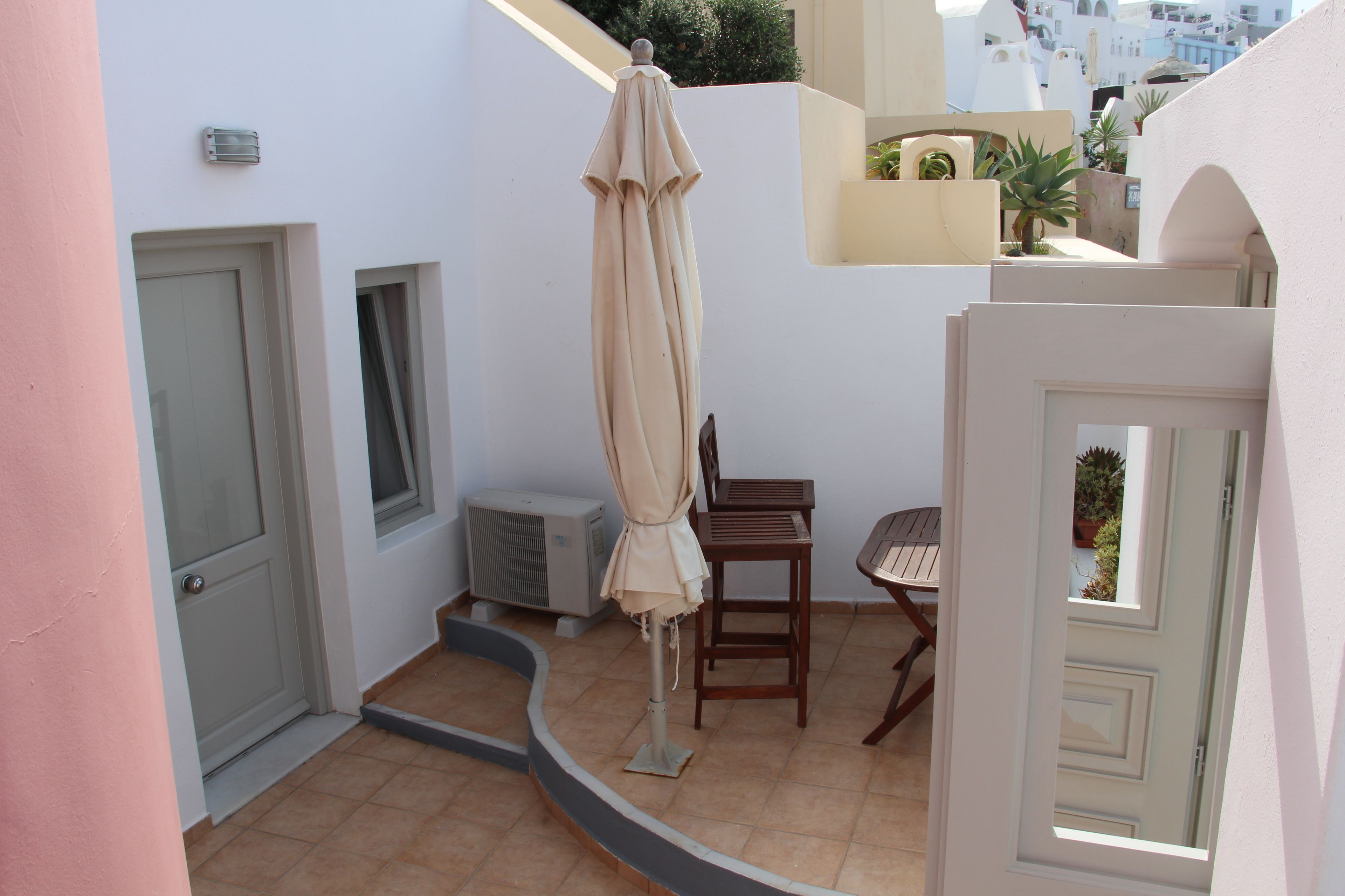 Un espacio exterior pequeño y cerrado con una puerta, un equipo de aire acondicionado, una silla de madera y un paraguas color beige.