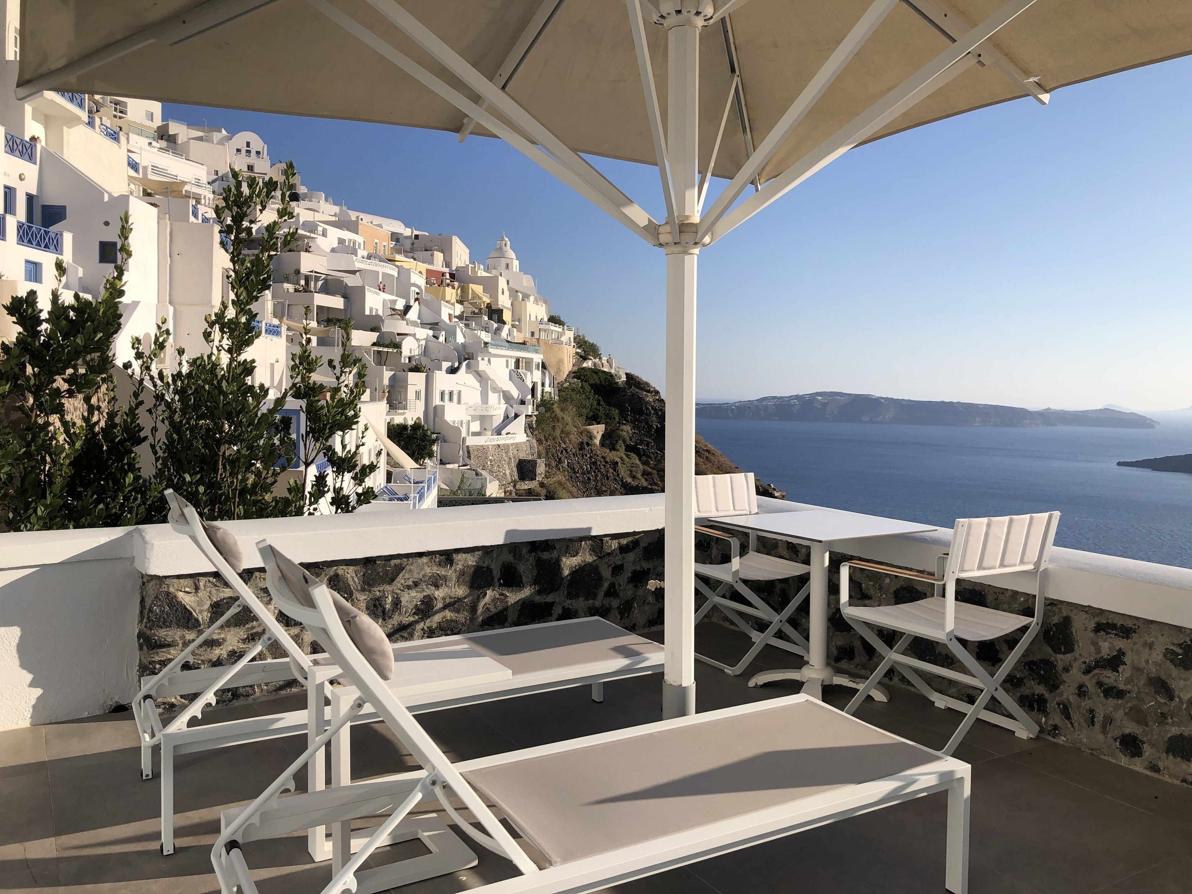 A seaside terrace with white lounge chairs and a table, overlooking a coastal town with white buildings and a view of the sea.