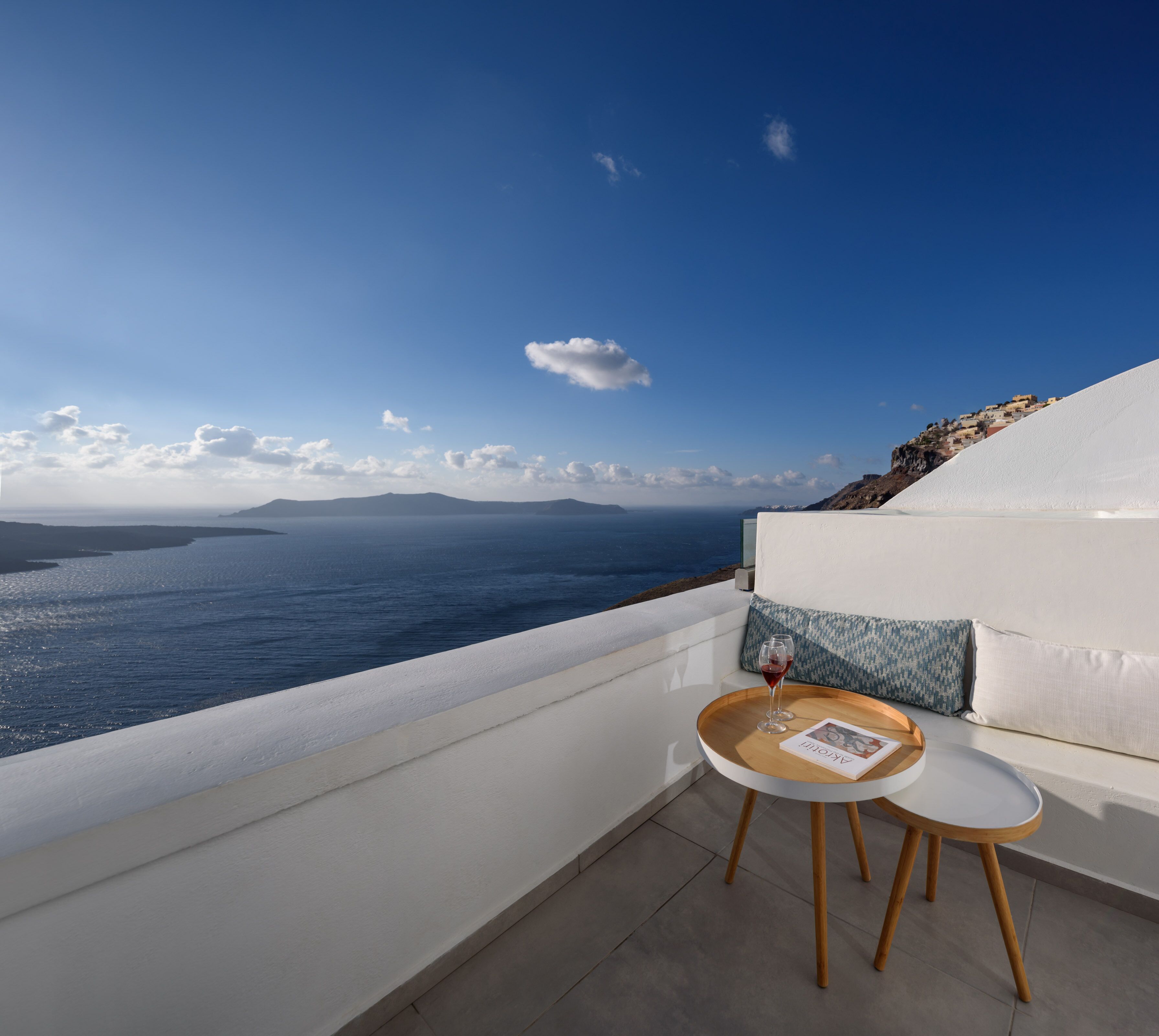 Ein Balkon mit weißer Reling und Blick auf das Meer, ein runder Tisch mit einem Glas Rotwein und ein Kissen mit geometrischem Muster.