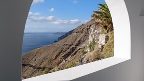 Vista a través de un ventanal arqueado blanco sobre un paisaje costero con acantilados rocosos y un cielo azul despejado.