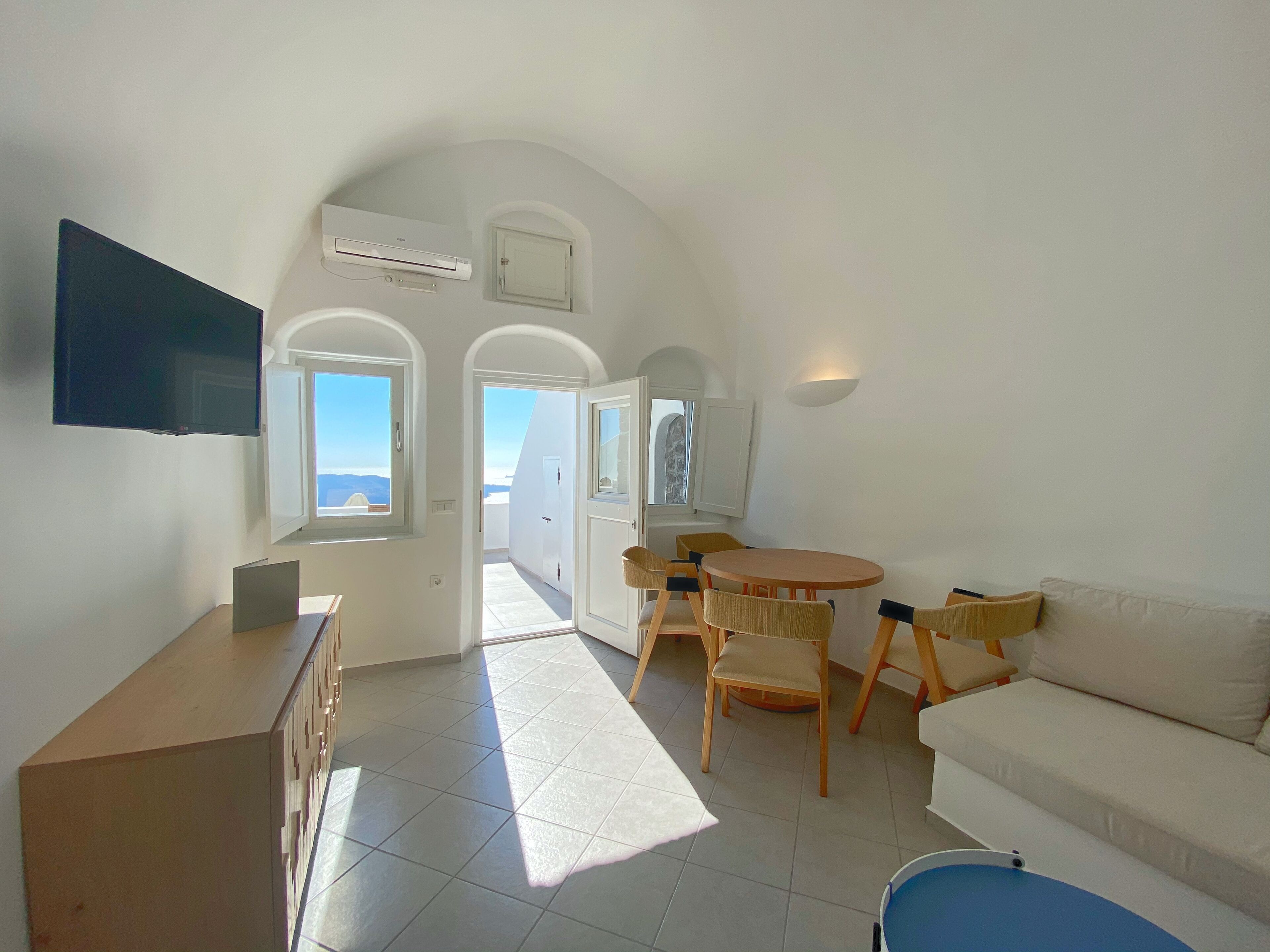 A modern, well-lit room with a flat-screen TV, a dining area with chairs and a table, a sofa, and a view through an arched doorway.