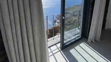 A room with a large sliding glass door leading to a balcony with a view of the ocean and hills.