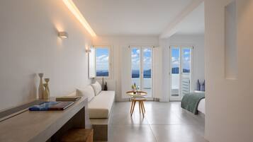 Une chambre d’hôtel moderne au design minimaliste, dotée d’une grande fenêtre offrant une vue sur la mer, d’une petite table ronde avec une bouteille et un verre, et d’un canapé recouvert d’un plaid vert.
