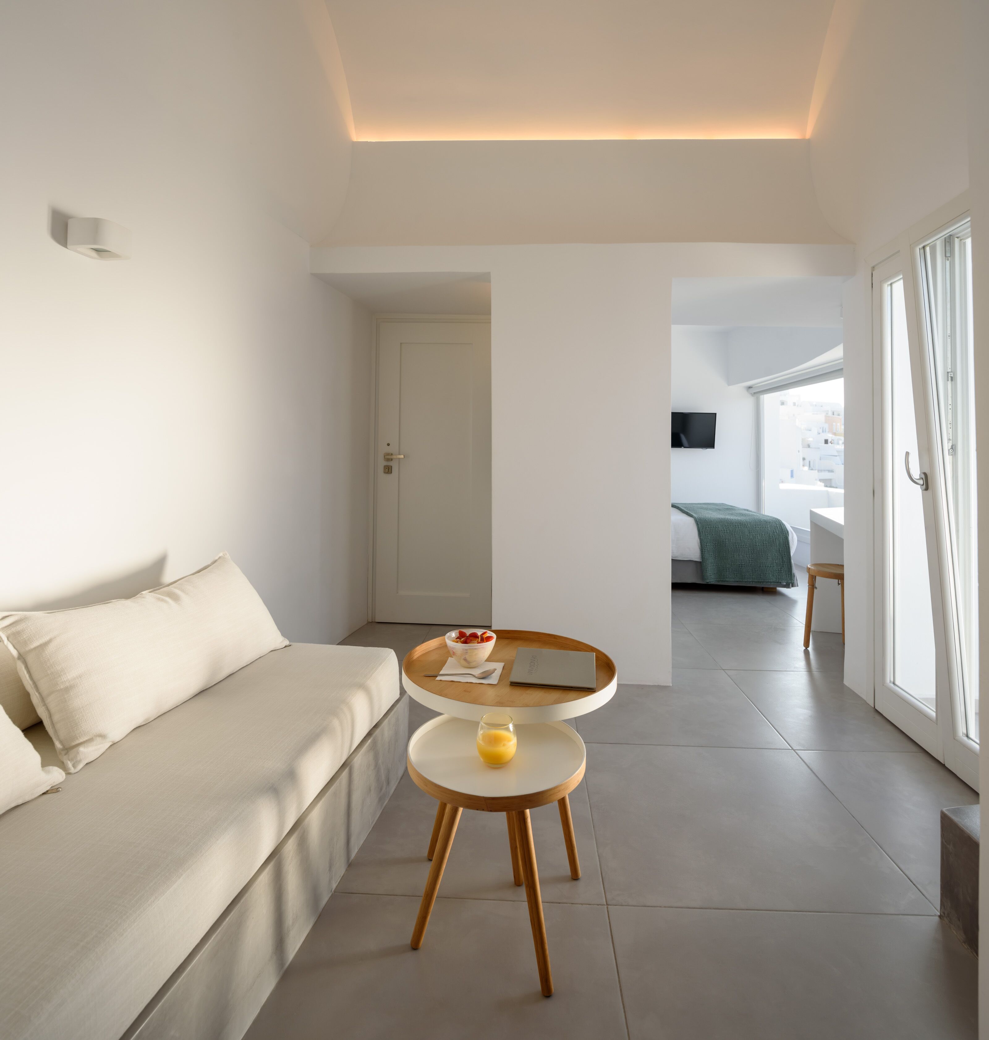 A modern, minimalist room with a sofa, a small round table with a bowl of fruit and a glass of orange juice, and a view of another room through an open door.