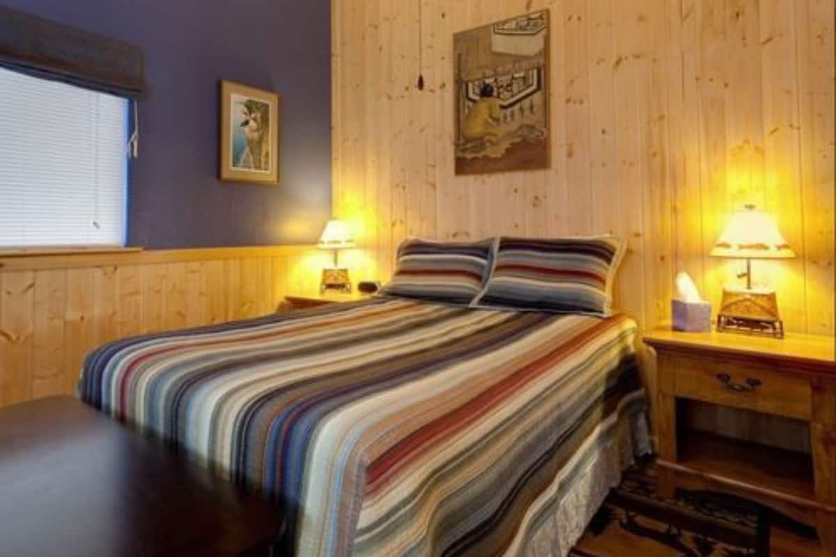 A cozy room with a striped bed, wooden walls, two bedside lamps, and a window with blinds.